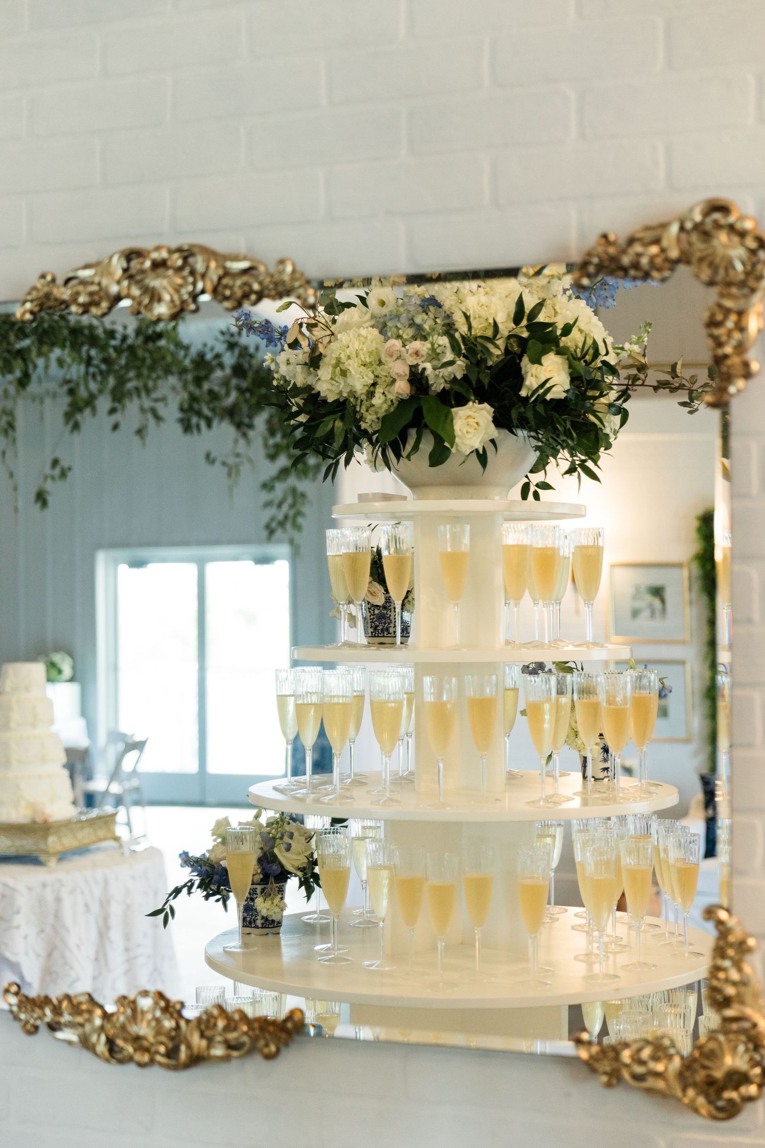 Elegant champagne tower with white floral arrangement reflected in ornate gold-framed mirror at wedding reception.