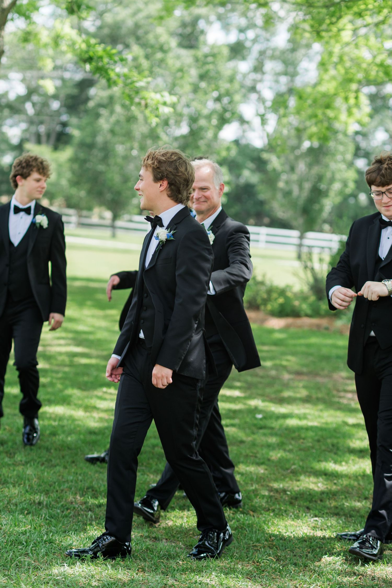Groomsmen in black tuxedos with bow ties walk outdoors on green grass at a wedding celebration.