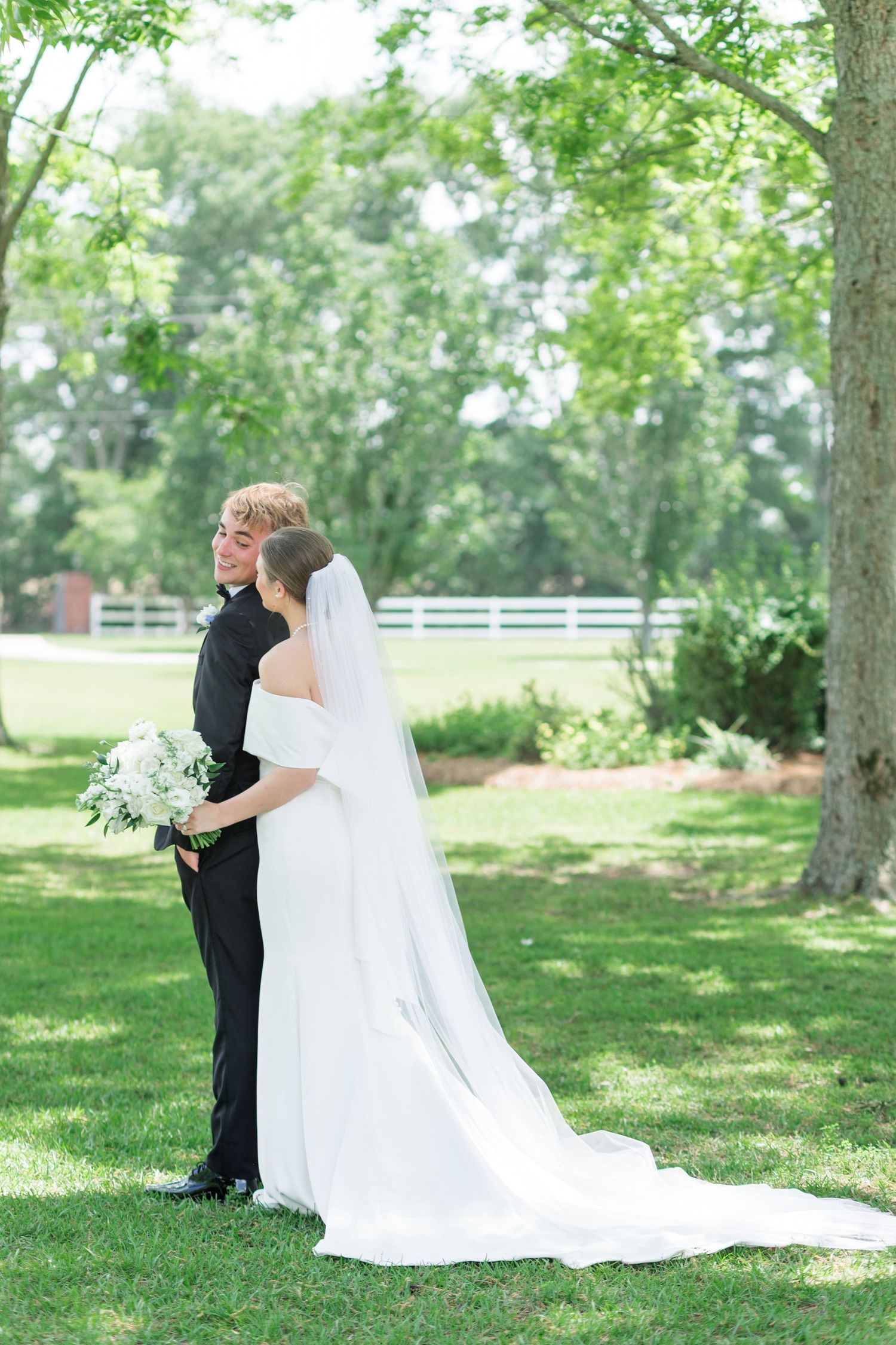Bride and groom embracing under lush green trees, bride wearing long white veil and gown.