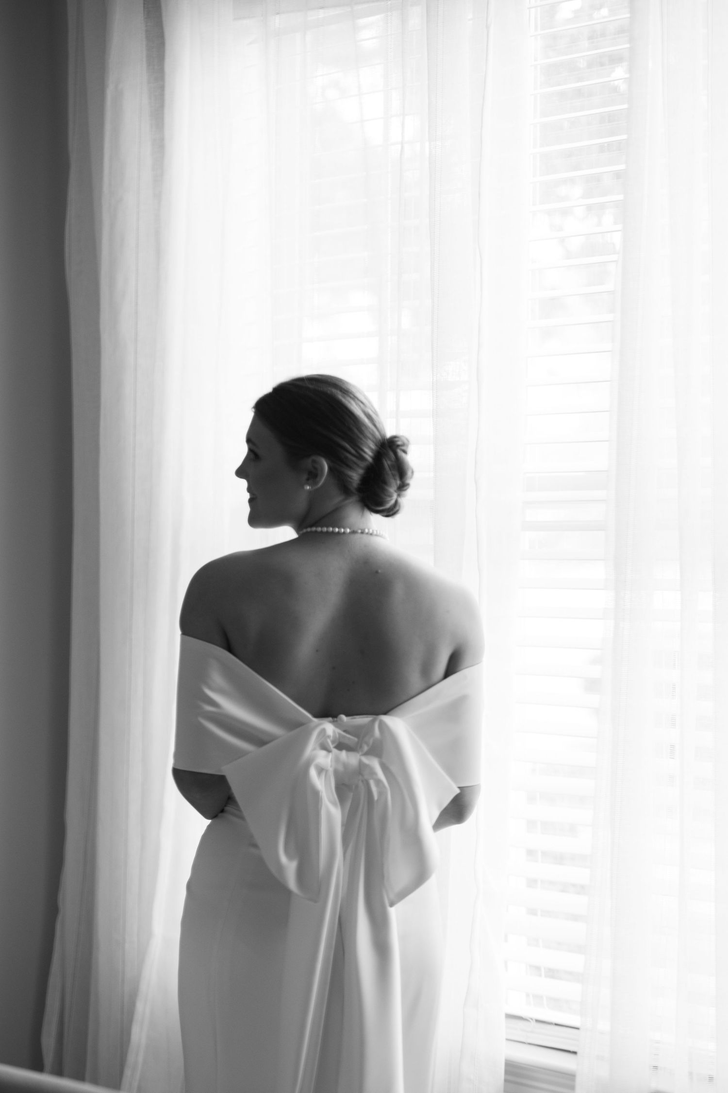 Elegant bride in an off-shoulder gown with a large bow stands before sheer curtains in a black and white photo.
