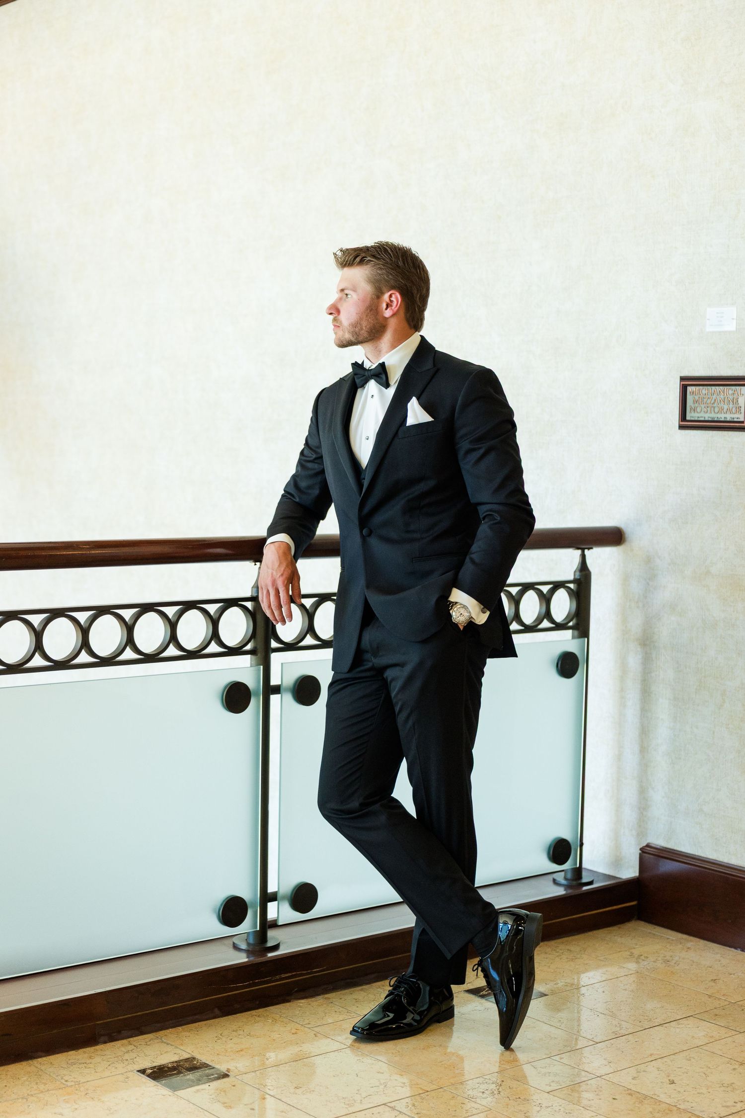 A dapper groom in a black tuxedo with bow tie leans against a glass railing in an elegant venue.