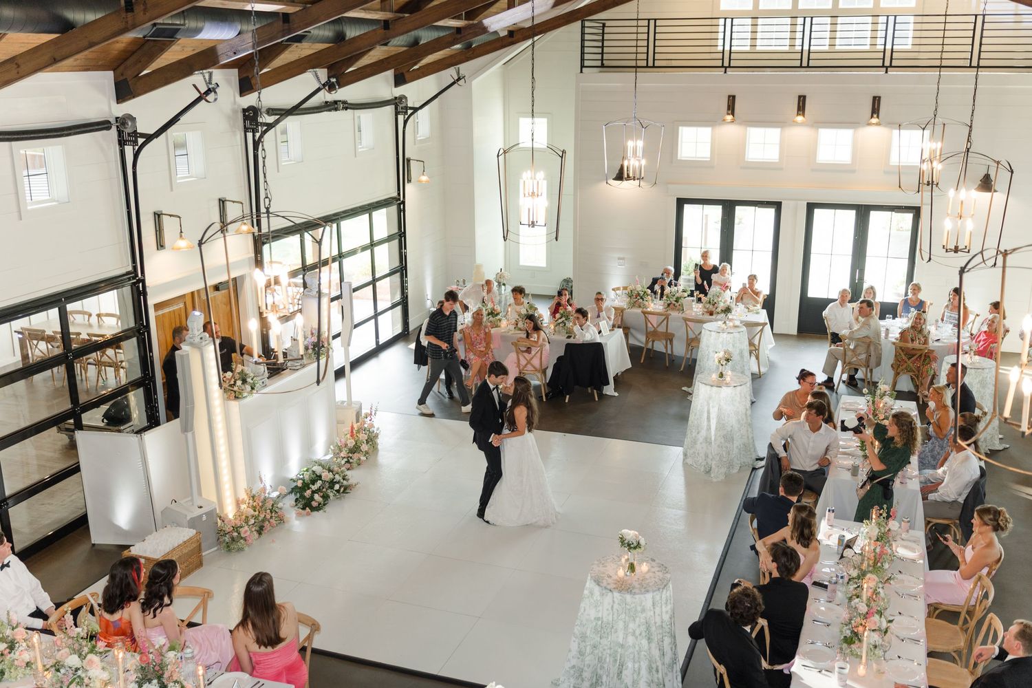 Elegant wedding reception in a bright barn venue with guests seated at decorated tables surrounding a white dance floor.