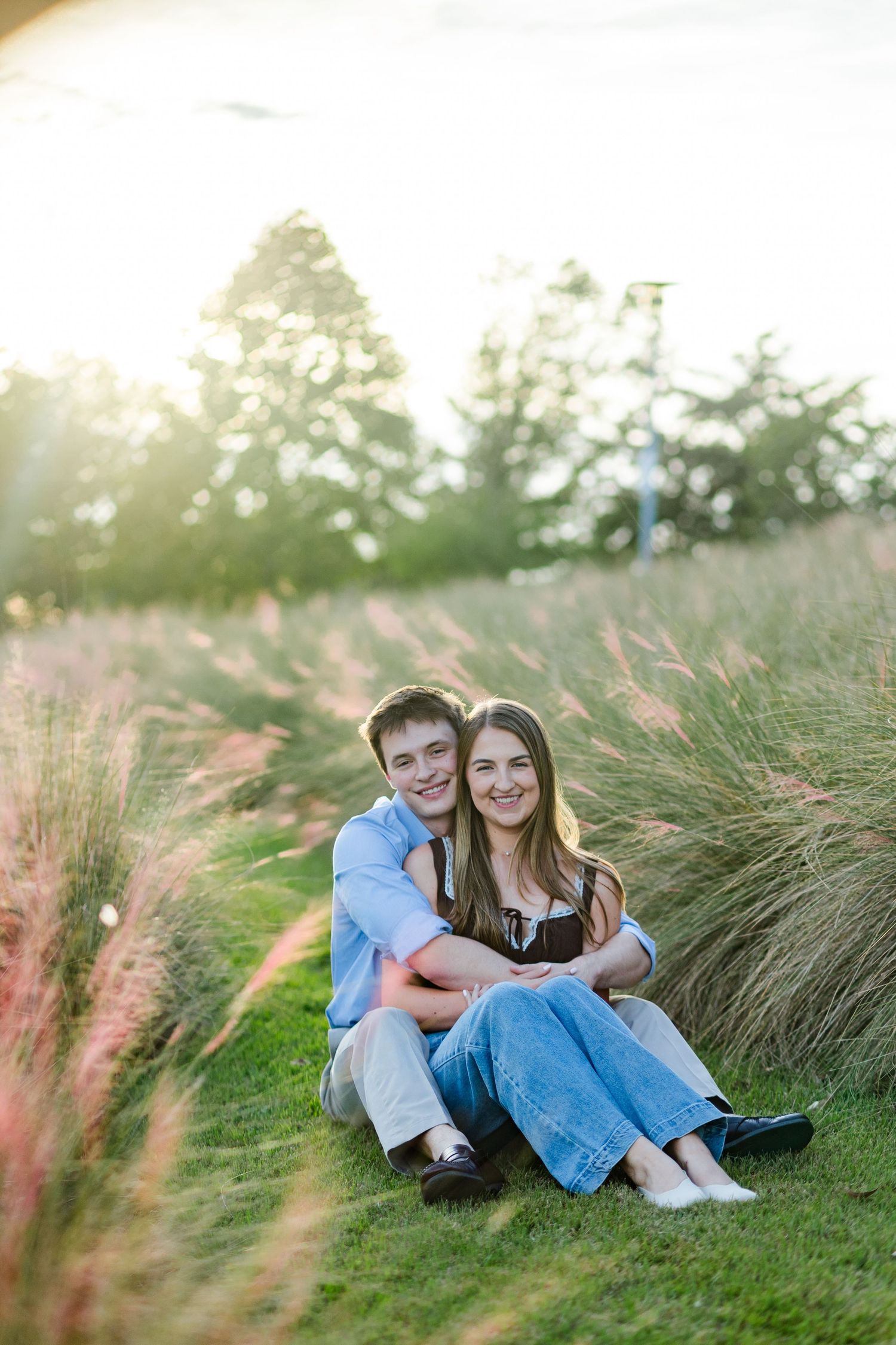 A happy couple sitting together on grass surrounded by tall ornamental grasses in golden sunlight.