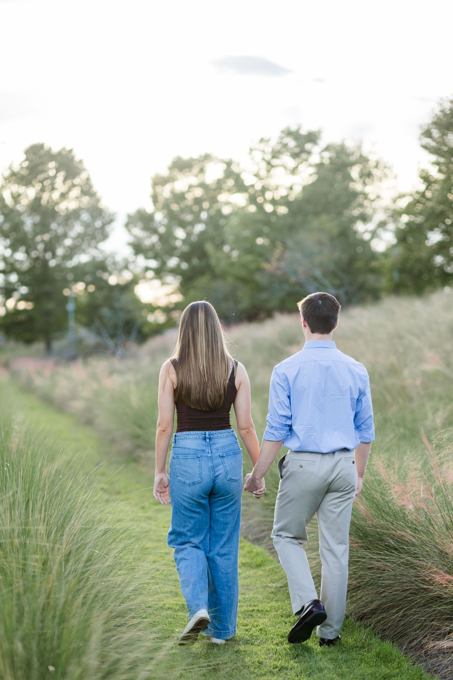 A couple holds hands while walking through a lush green garden path surrounded by tall grasses and trees at sunset.