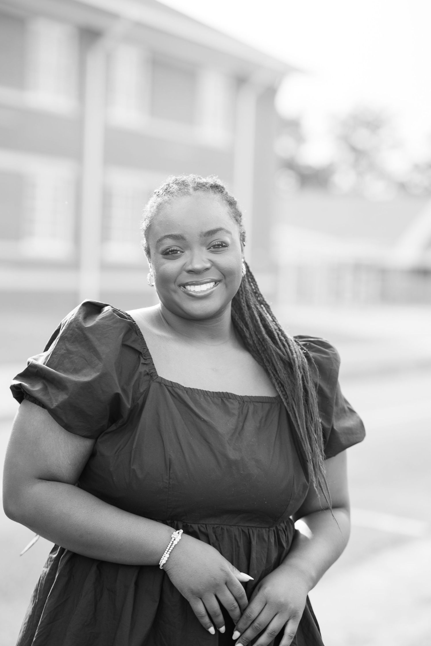 Black and white portrait of a smiling woman in a puff-sleeve dress, posing outdoors near a building.