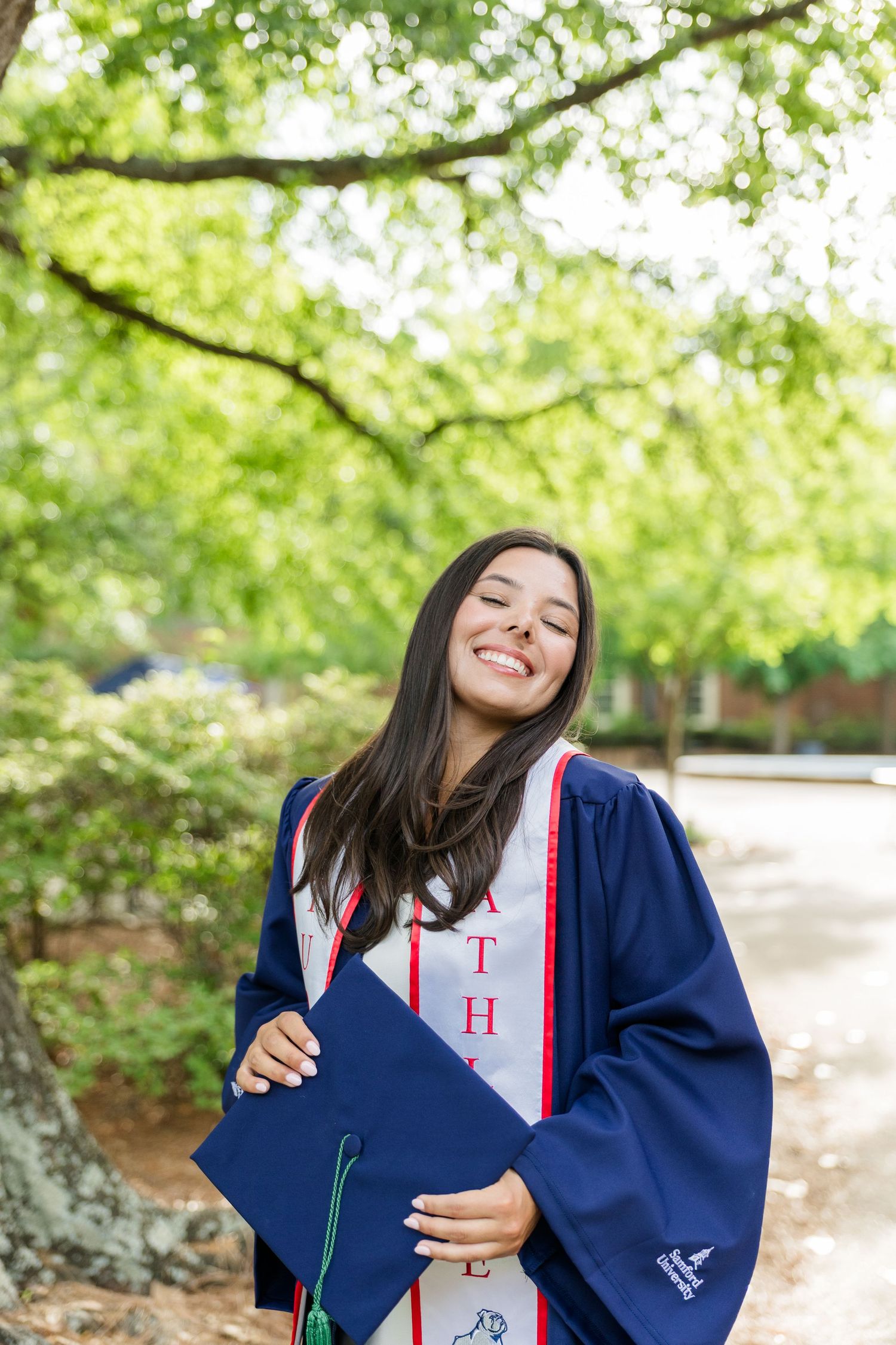 Smiling female graduate in navy blue cap and gown holding mortarboard outdoors under green trees.