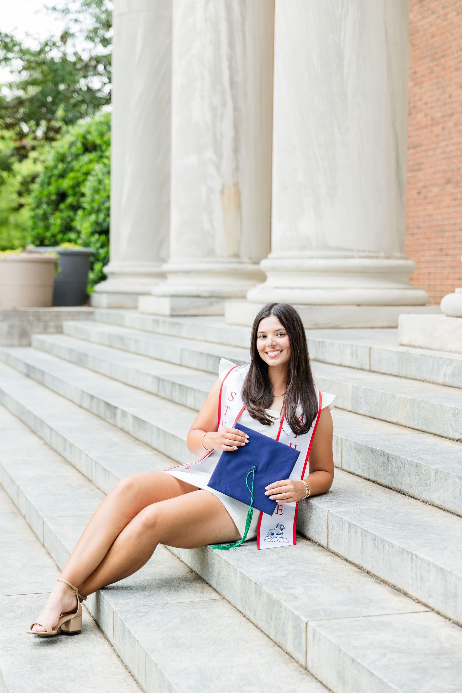 Smiling graduate sits on university steps holding cap and diploma, wearing white dress and academic regalia.