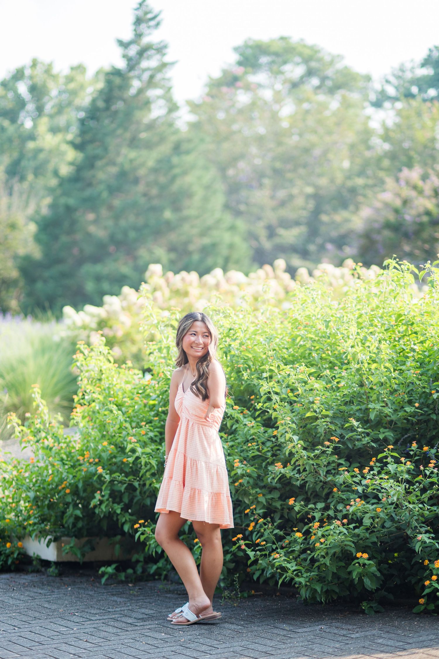A young woman in a peach dress smiles while standing beside lush green flowering garden shrubs.