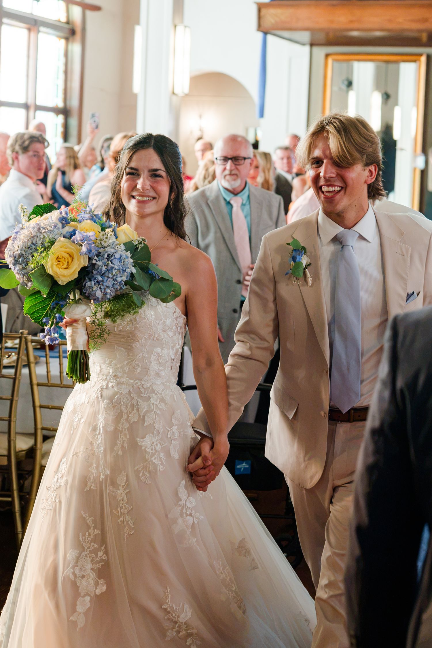Joyful newlyweds walking down aisle, bride holding blue and yellow bouquet in lace wedding gown.