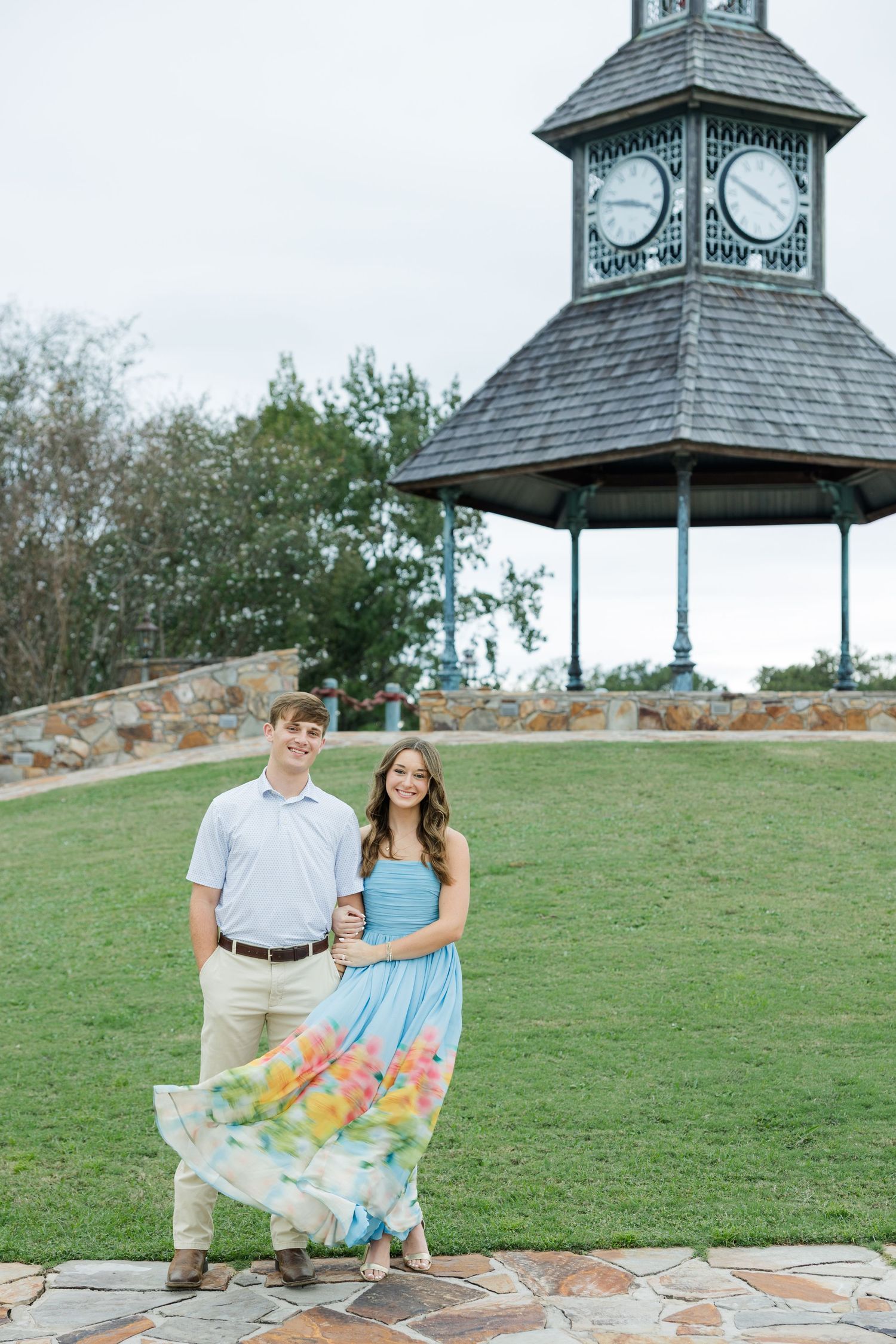 Couple stands arm in arm on a green lawn in front of a stone clock tower gazebo during an engagement photo session.