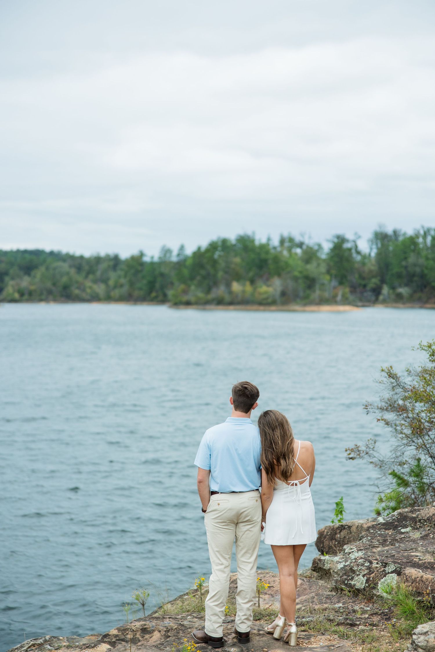 Couple standing close together on a rocky lakeside outcrop, gazing out over the water toward a forested shoreline.