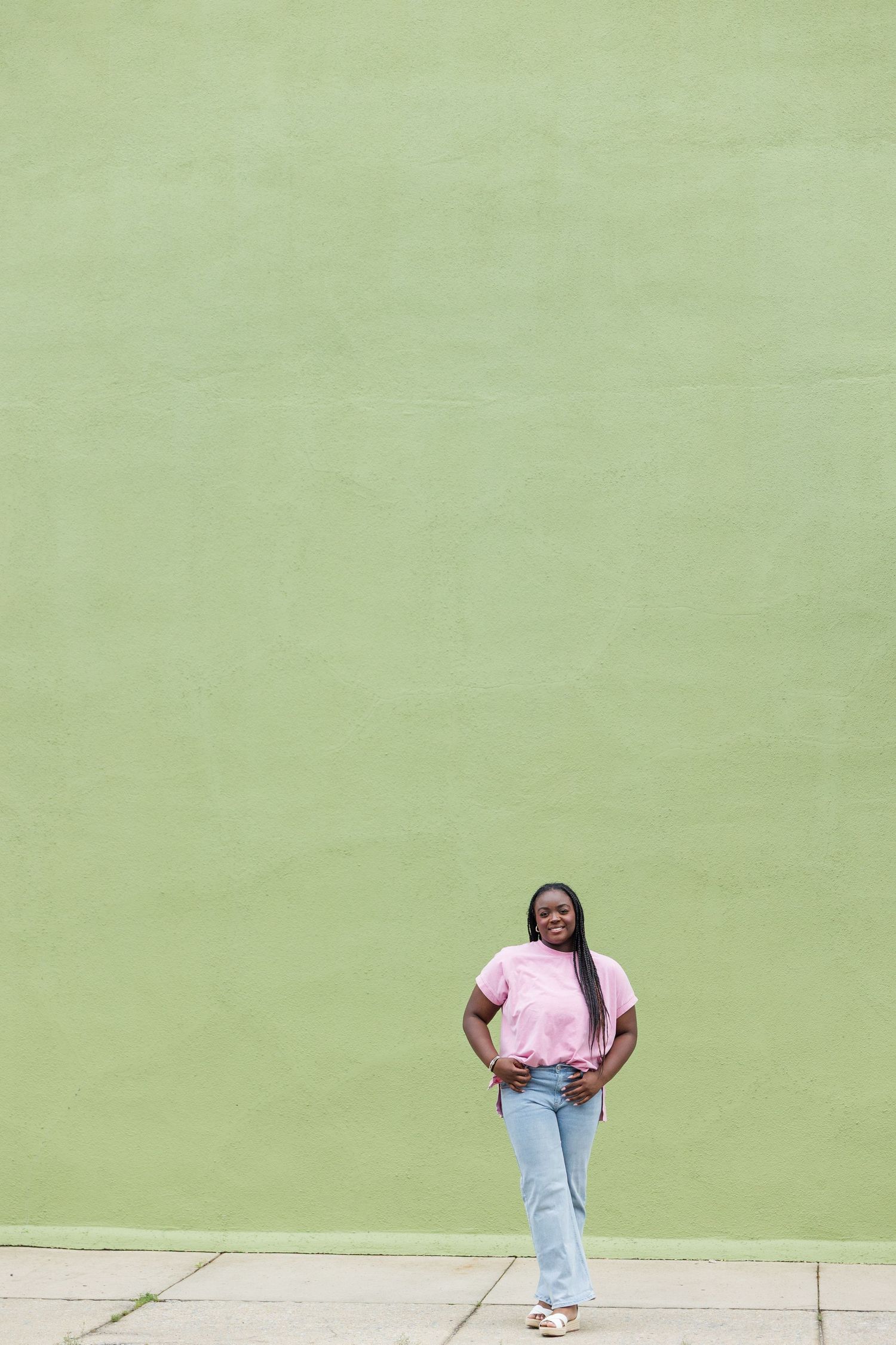 Young Black woman in pink shirt and jeans standing against green wall outdoors.