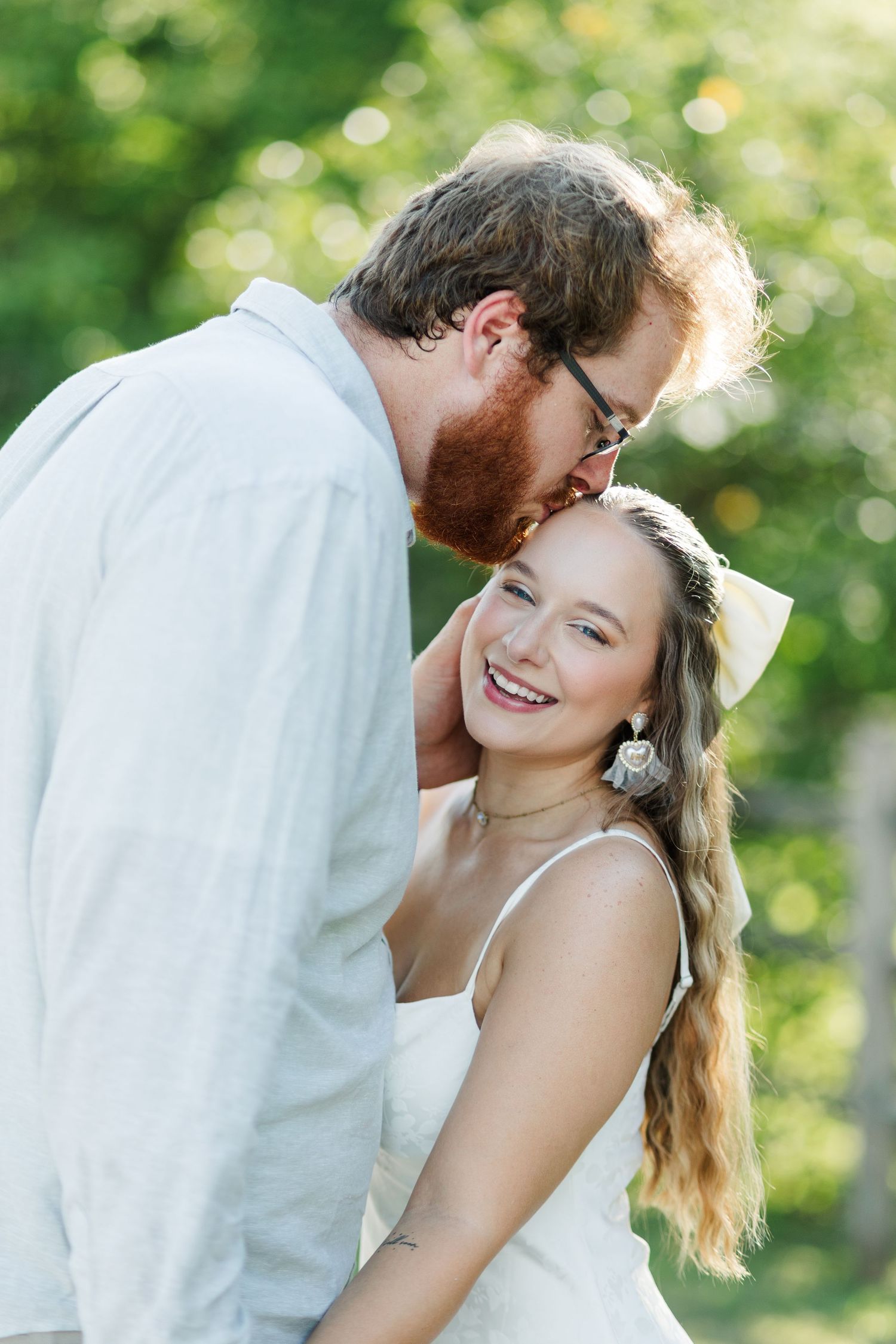 A bearded groom in white kisses his smiling bride wearing a white bow and dress outdoors.