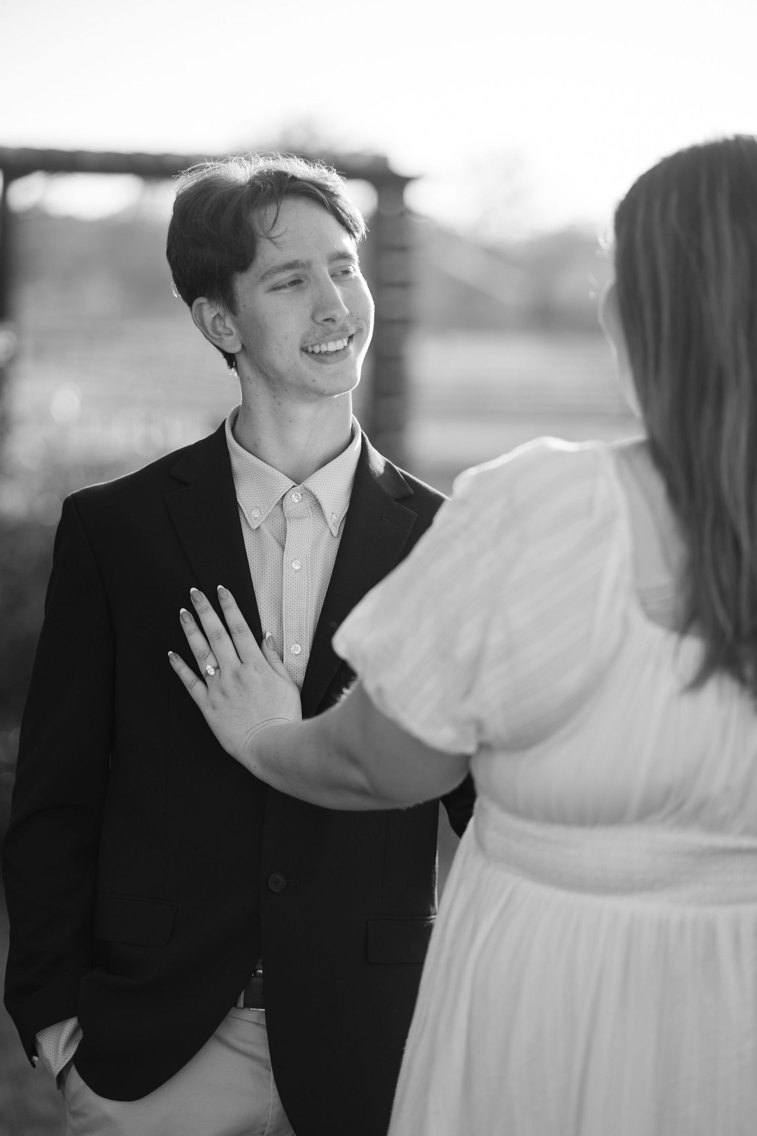 Black and white photo of a smiling groom in suit as bride in white dress shows off her engagement ring.