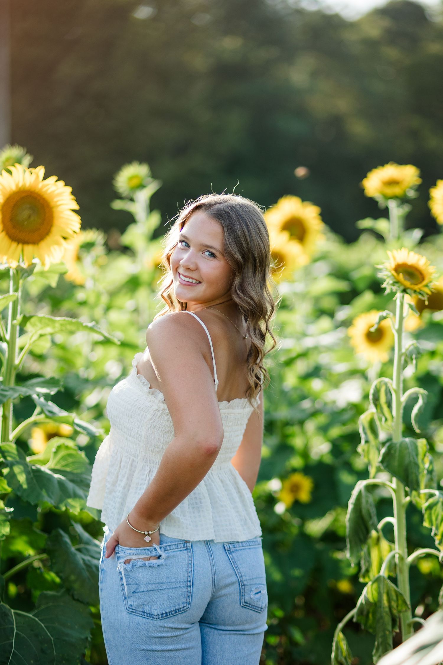 Smiling young woman in white top and jeans standing among tall sunflowers in a sunny field.