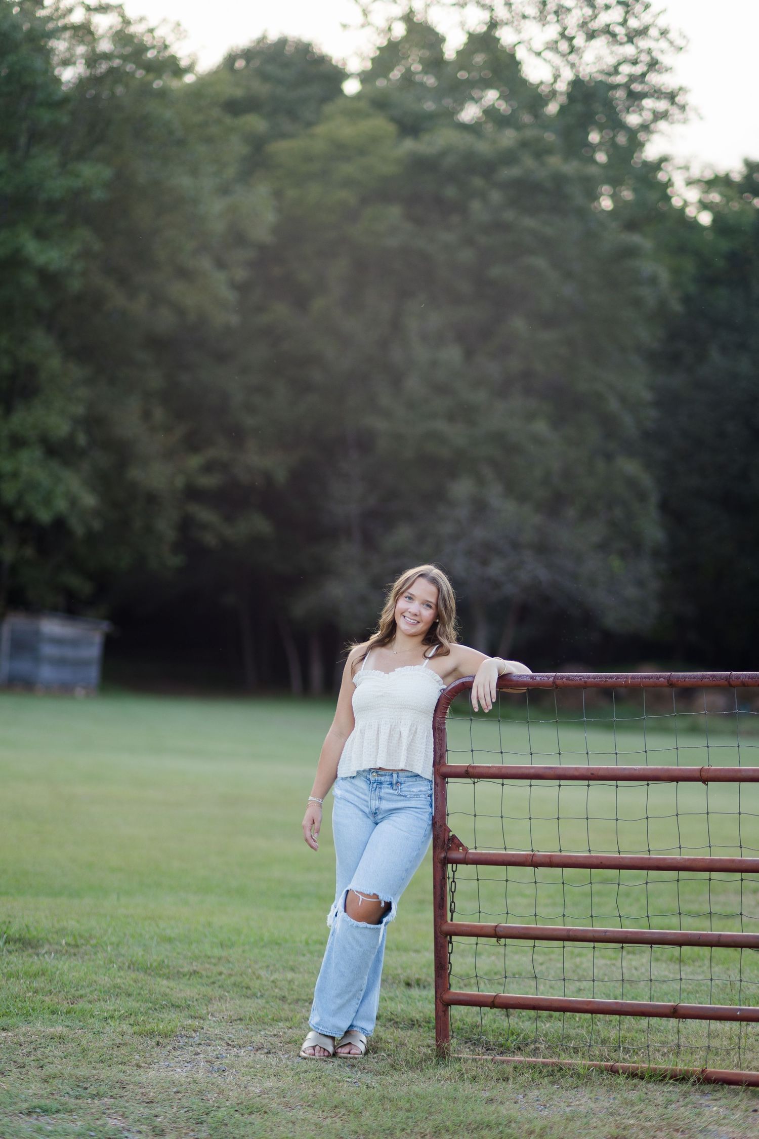 Young woman in white top and ripped jeans leaning on a red farm gate in a green field.