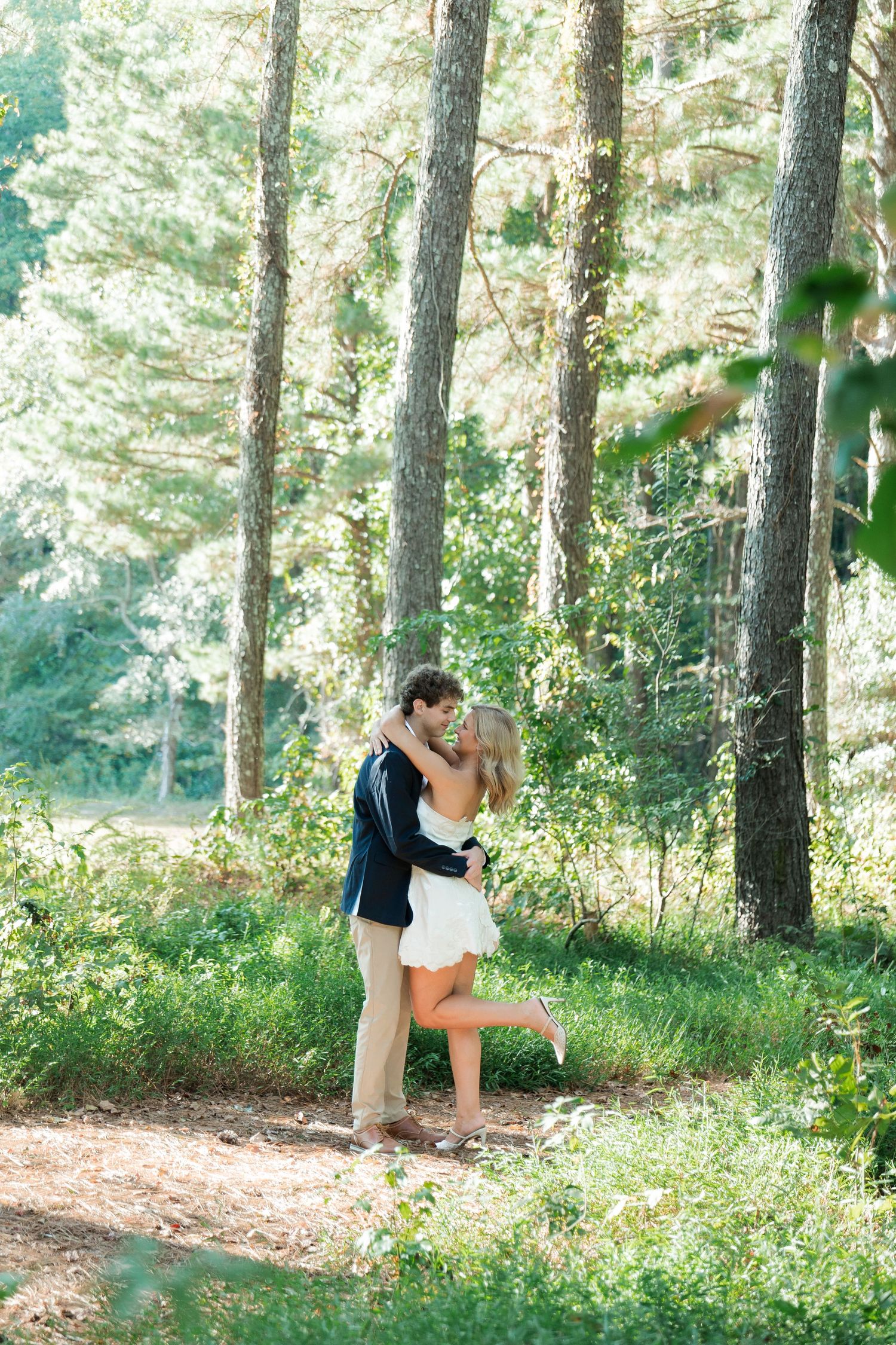 Couple sharing romantic kiss among tall pine trees in sunlit forest clearing.