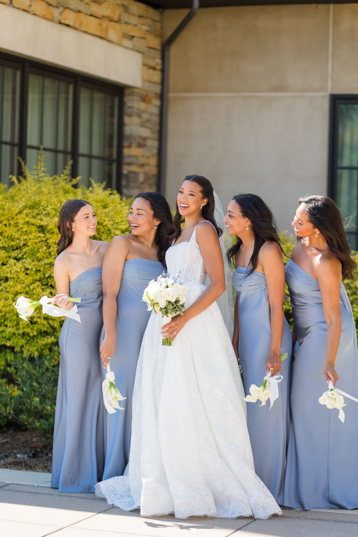 Bride in white gown laughing with four bridesmaids in dusty blue dresses holding white bouquets outdoors.