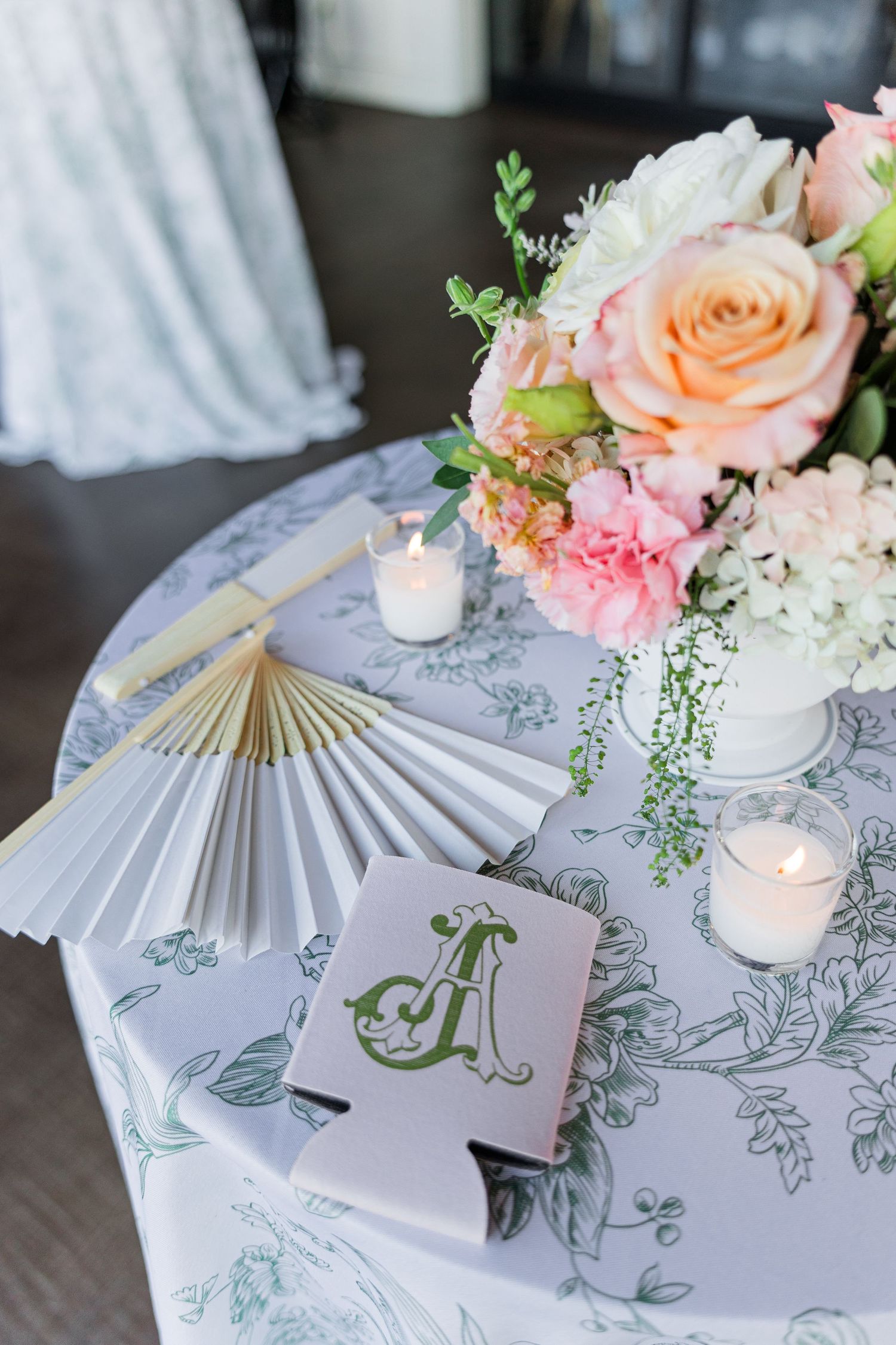 Elegant wedding table with floral centerpiece, paper fans, monogrammed koozie, and candles on floral tablecloth.