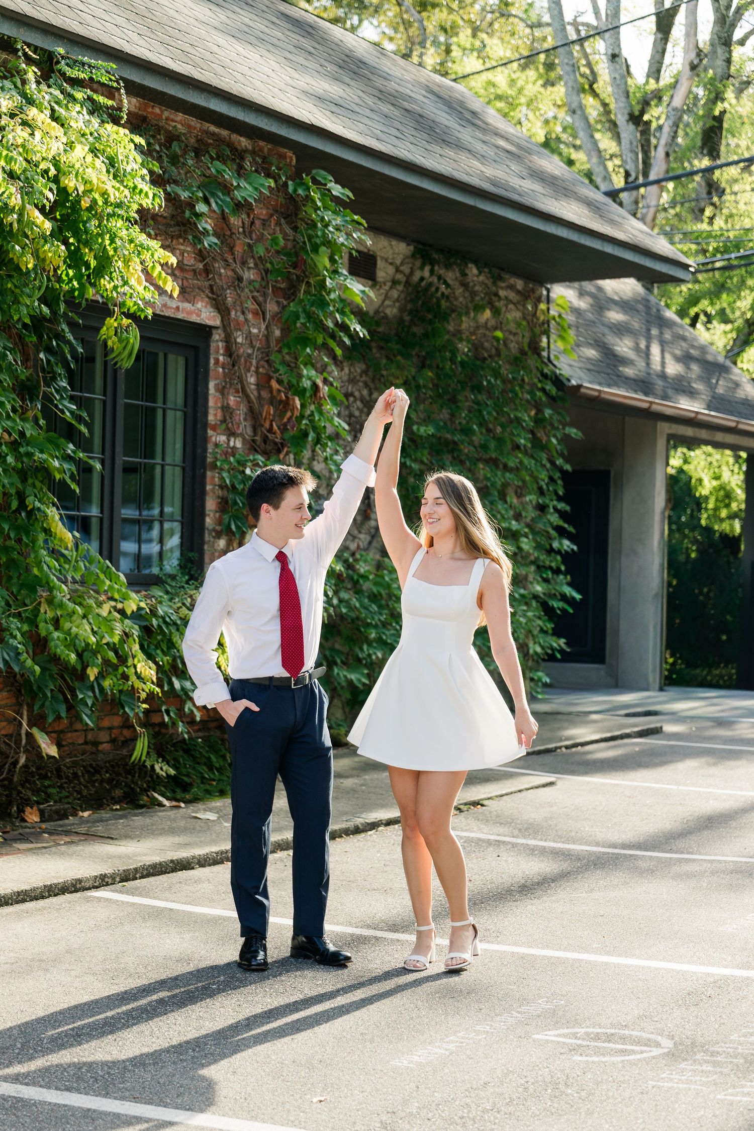 A couple dances in a parking lot, man in white shirt and red tie holds woman in white mini dress hand up.