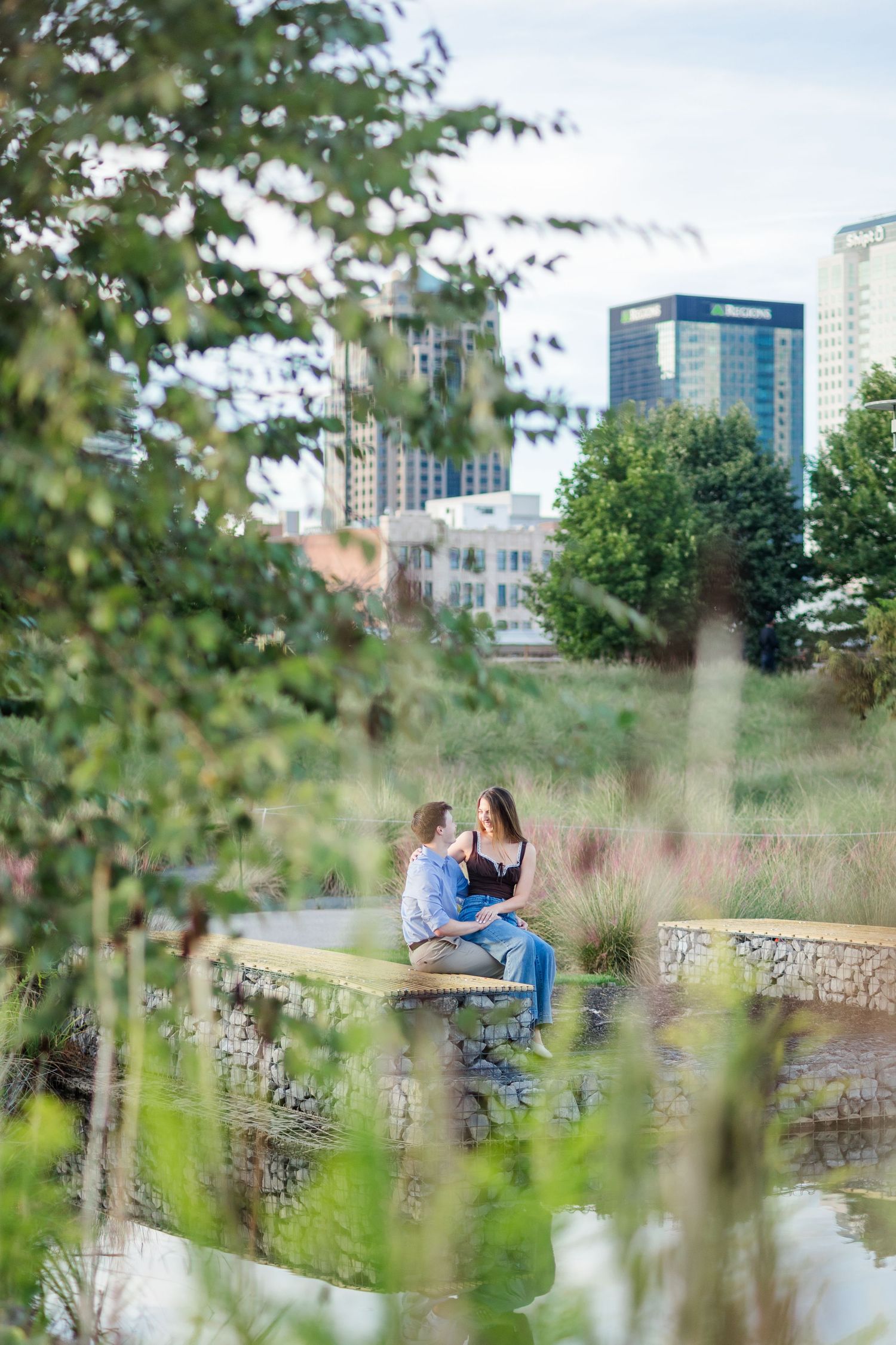 A couple sits on a stone ledge in an urban garden park with a city skyline visible in the background.