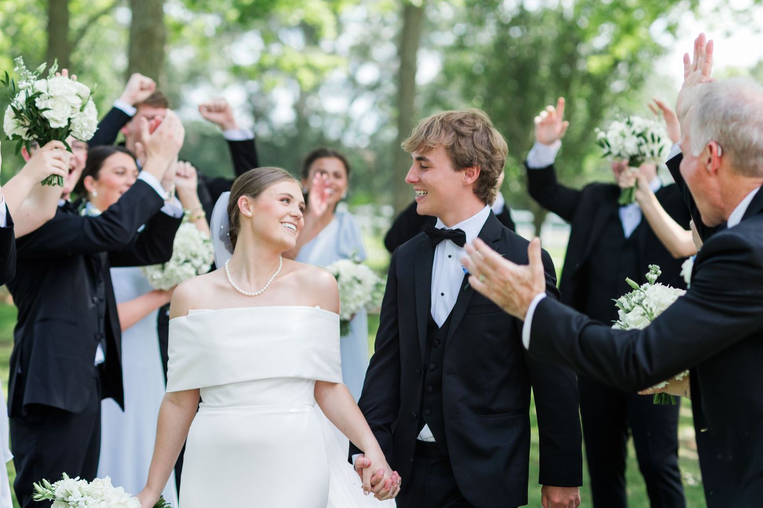 Joyful newlyweds walk hand in hand as wedding party celebrates with raised bouquets outdoors.