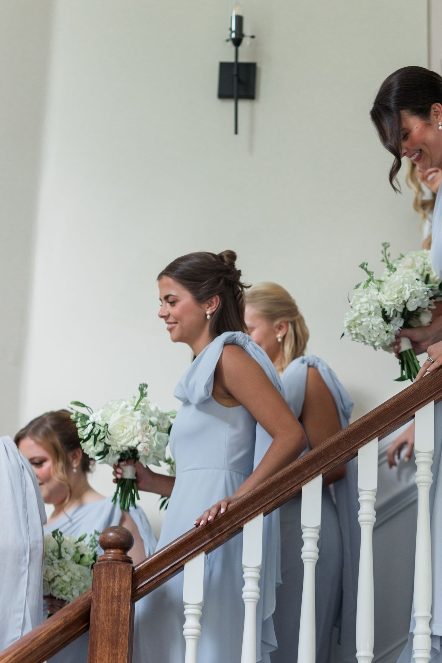 Bridesmaids in light blue dresses descend staircase holding white hydrangea bouquets at wedding.