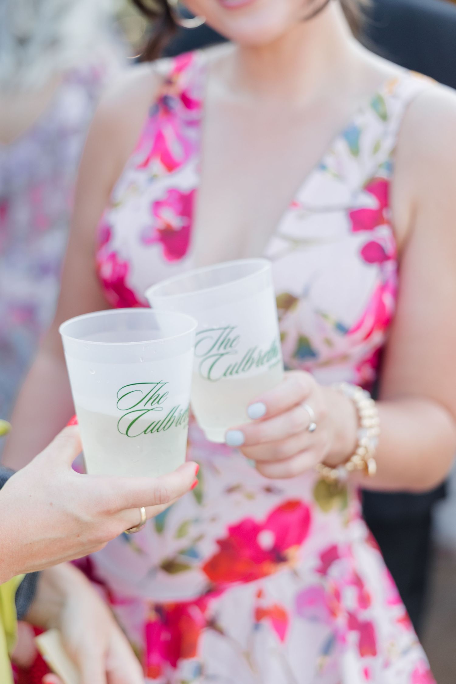 Two frosted cups printed with The Culbreth toasting at an outdoor event, held by a woman in a floral dress.