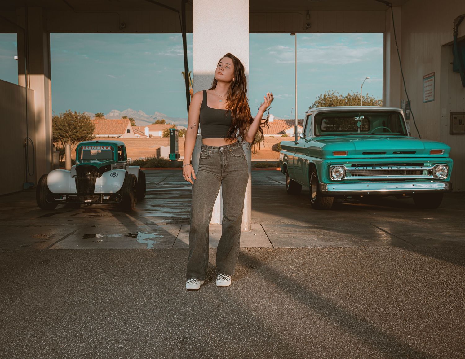 Young woman holding a guitar poses between vintage cars at a gas station, wearing crop top and flared jeans.
