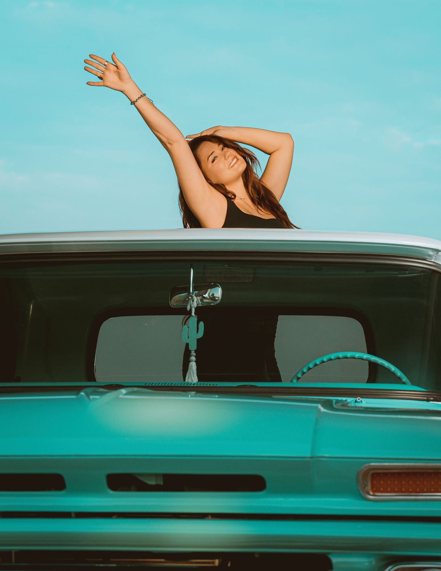 A young woman stretches her arms up joyfully through the sunroof of a vintage turquoise truck under a blue sky.