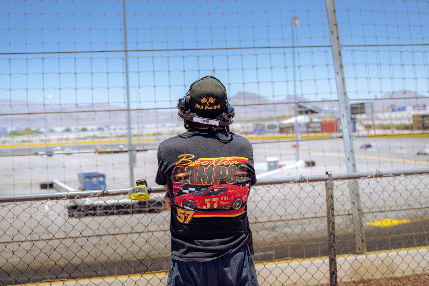 Person in racing shirt watches cars on a racetrack through a chain-link fence on a sunny day.