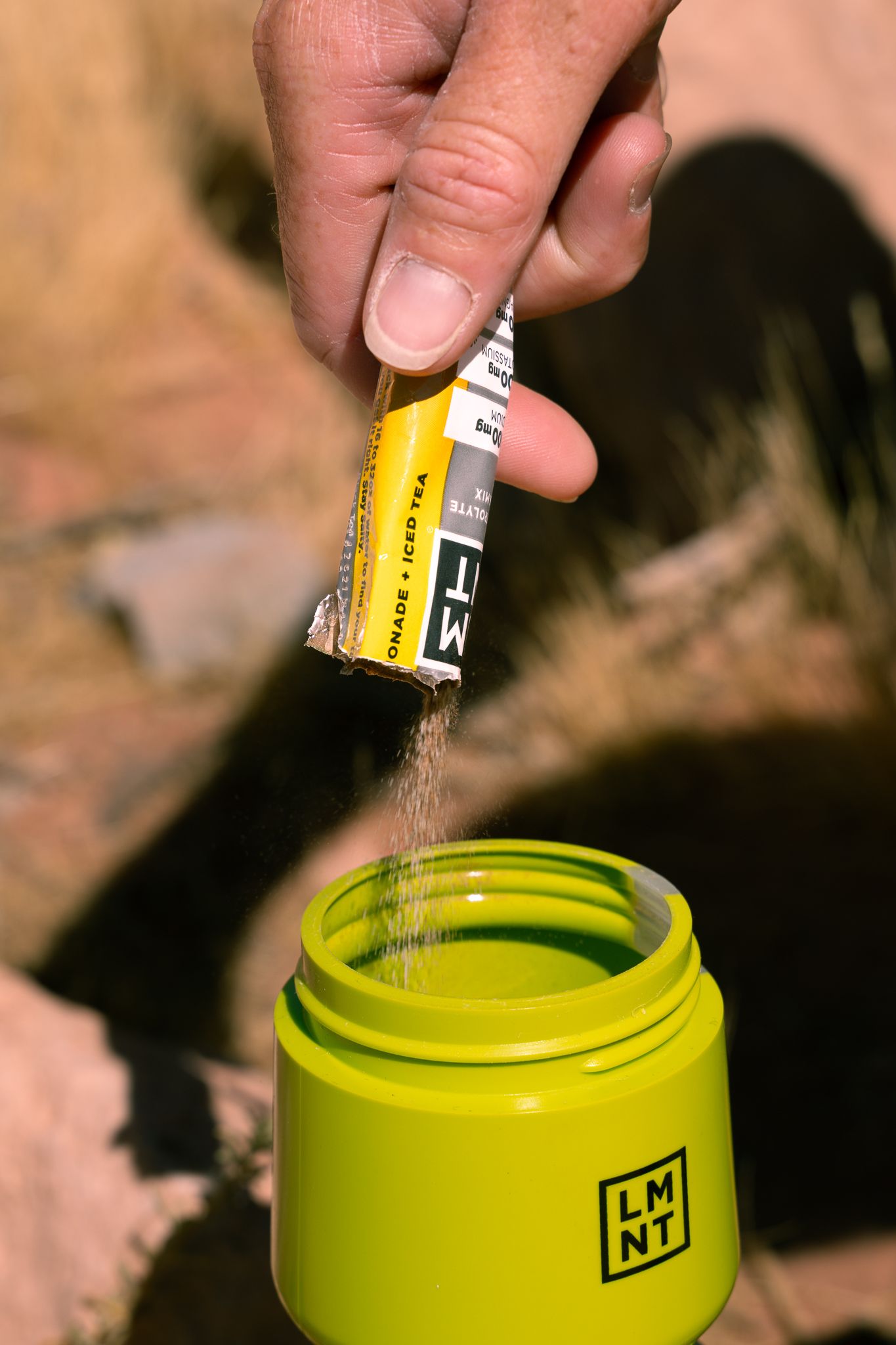 Hand pouring LMNT lemonade iced tea electrolyte powder packet into yellow water bottle outdoors.