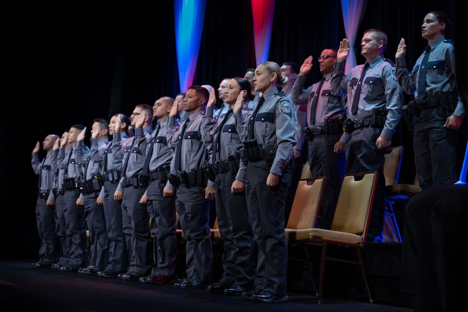 Police academy graduates in uniform raise right hands taking oath of service on stage at formal graduation ceremony.
