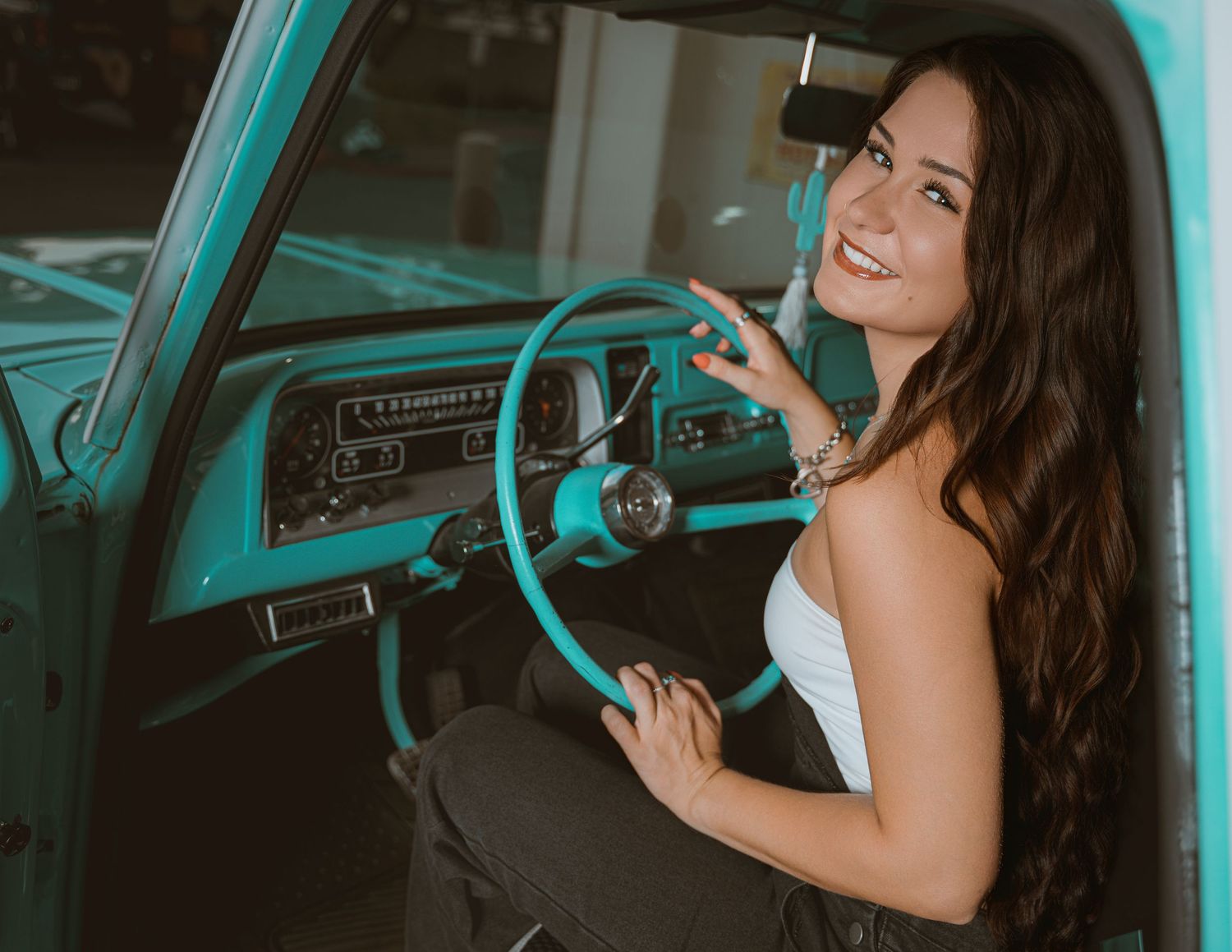 Young woman with long dark hair smiling in vintage teal truck, wearing white top, posing at steering wheel.