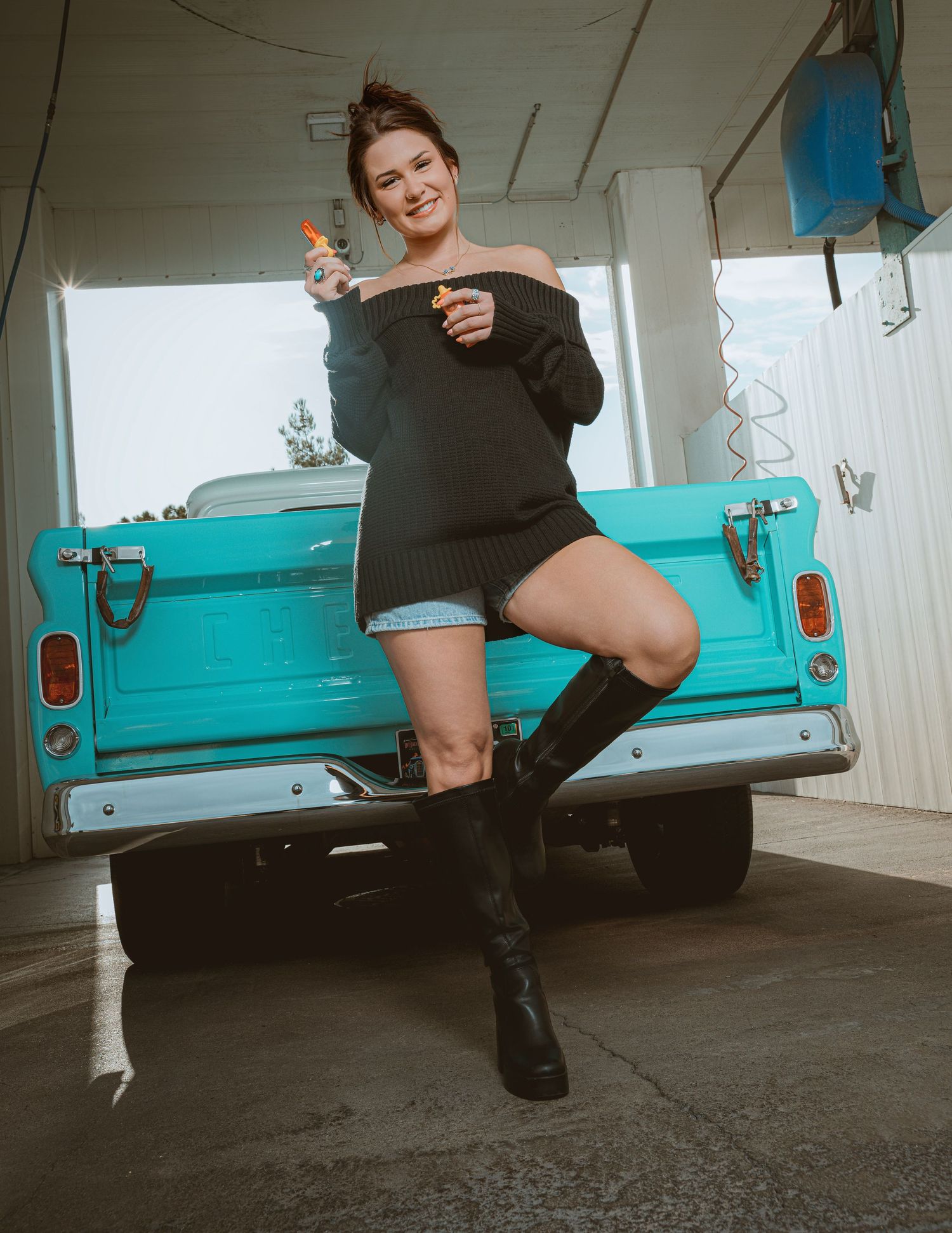 Young woman in black off-shoulder sweater and denim shorts posing playfully against a teal vintage pickup truck.