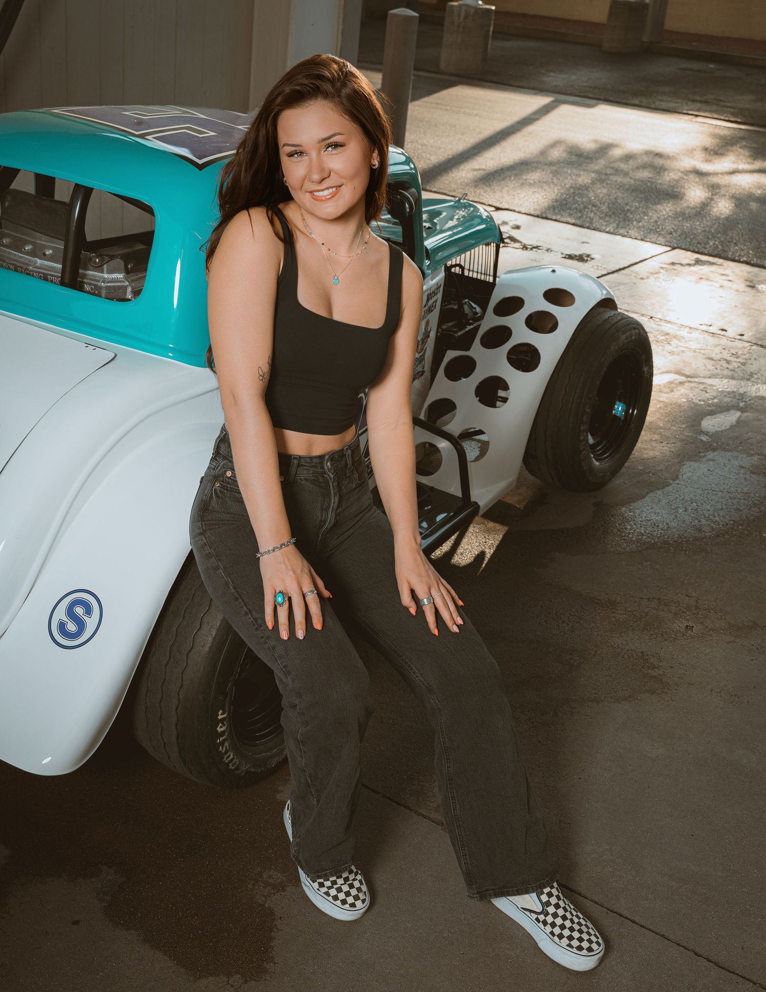 Young woman in black crop top and jeans posing with a teal and white vintage legend race car in a garage.