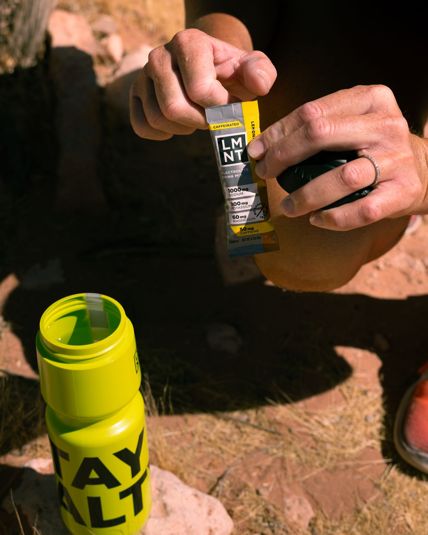 Hands opening an LMNT electrolyte packet above a yellow STAY SALTY bottle on rocky desert terrain.
