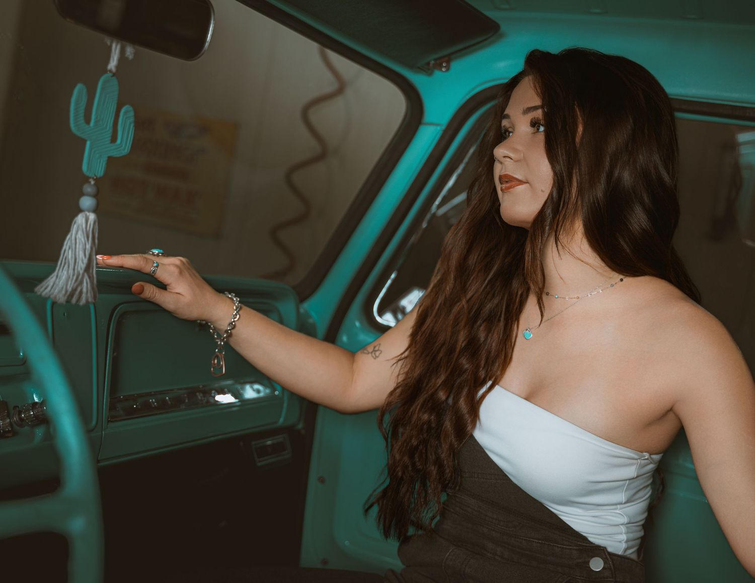 Young woman with long dark hair in a white tube top reaches for a cactus tassel charm inside a vintage teal truck.