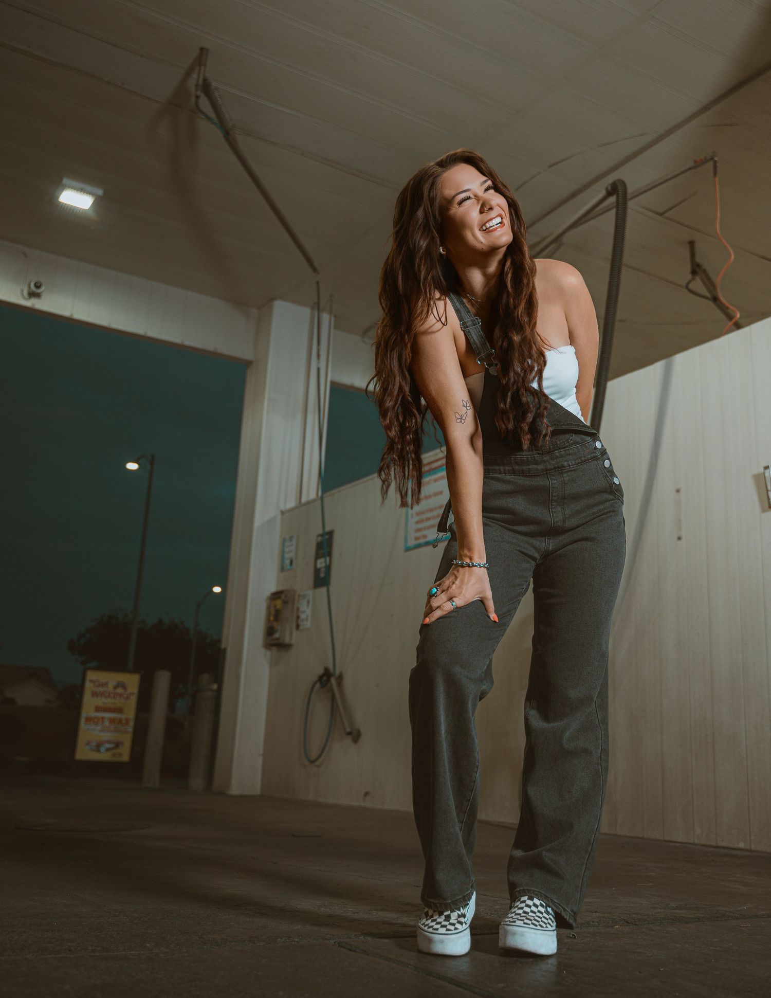Smiling young woman with long wavy hair wearing white top and dark jeans posing in a garage setting.