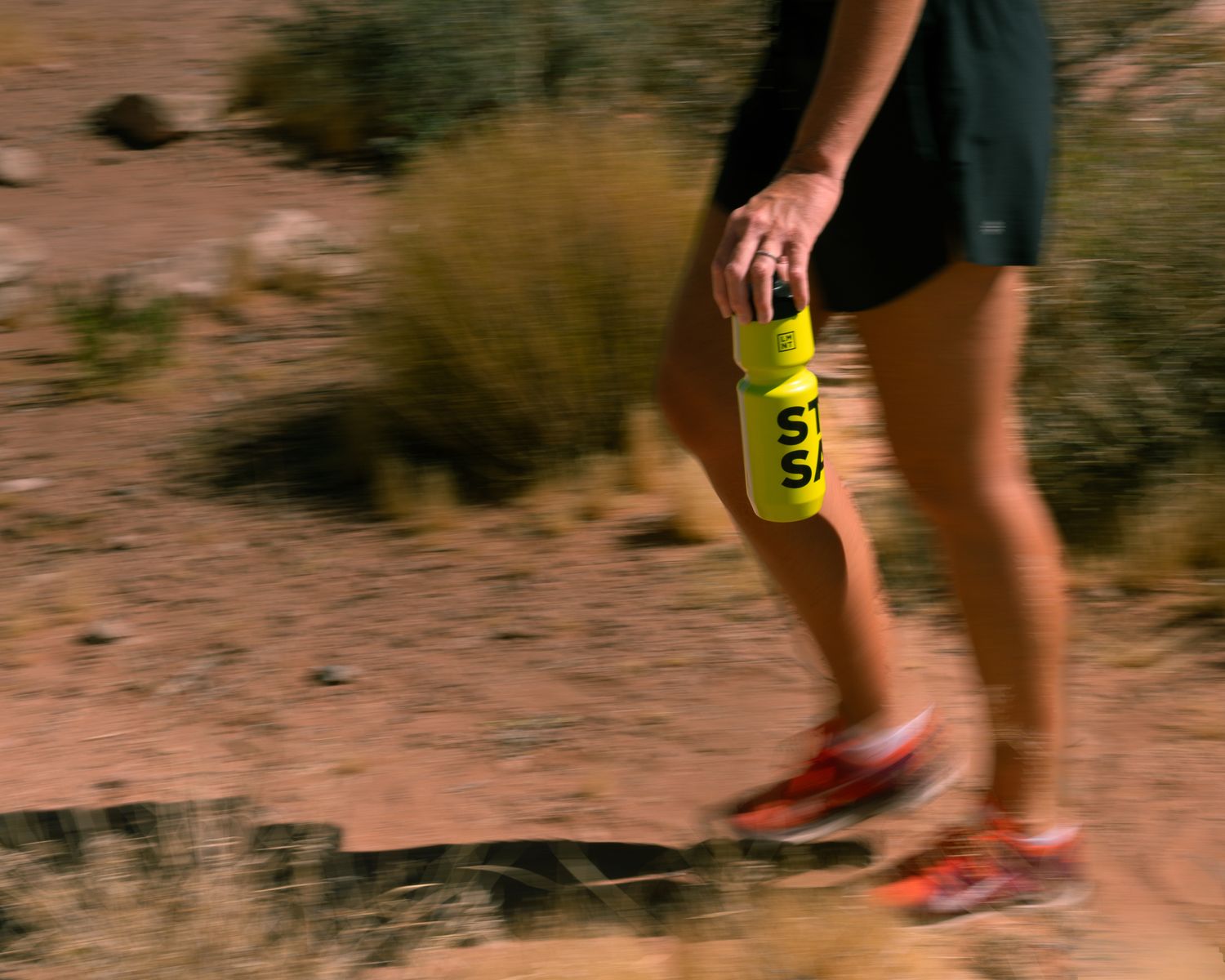 Female runner carrying a neon yellow LMNT sports bottle while jogging on a red dirt desert trail.