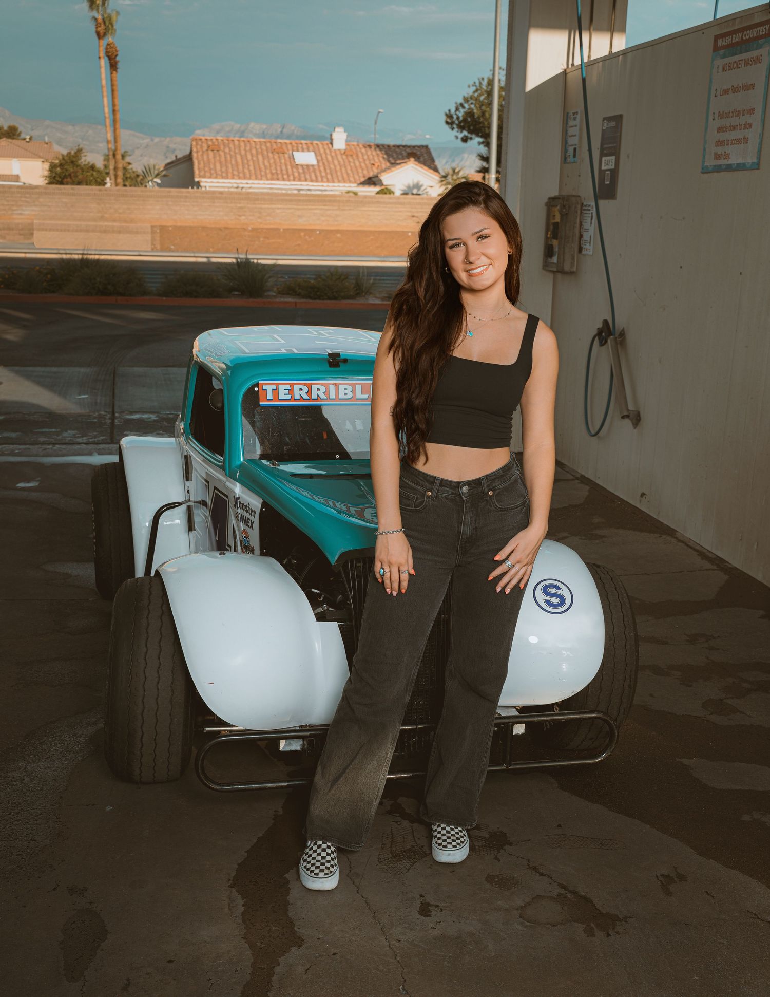 Smiling young woman in black crop top and dark jeans standing beside a teal vintage legend race car outdoors.