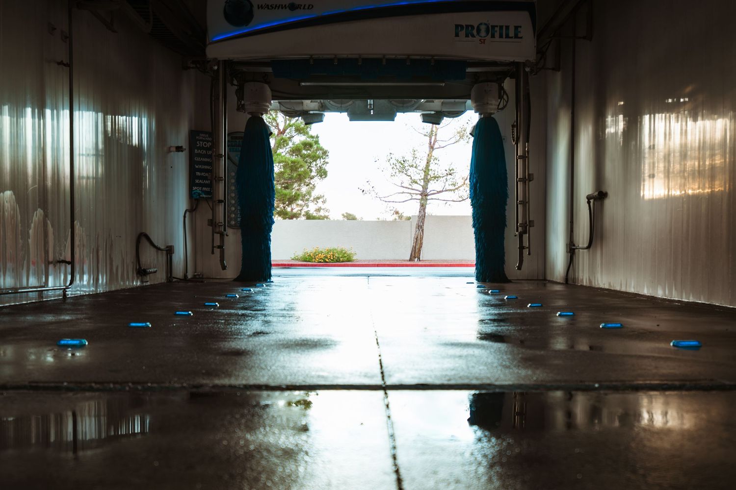 Interior view of a wet car wash tunnel with blue LED guides and open exit showing a sunny day outside.