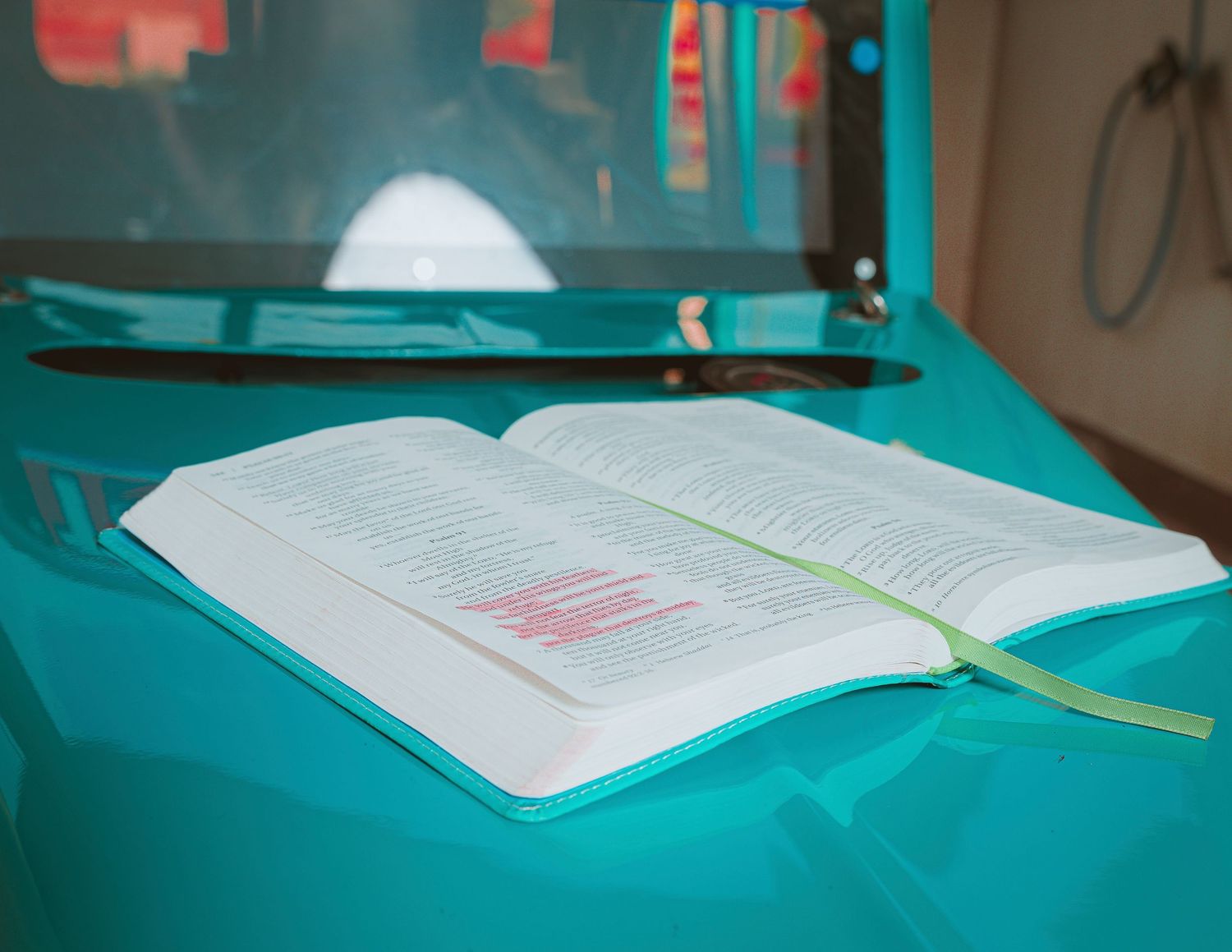 Open Bible with pink highlighted text resting on a teal cloth surface near a monitor.