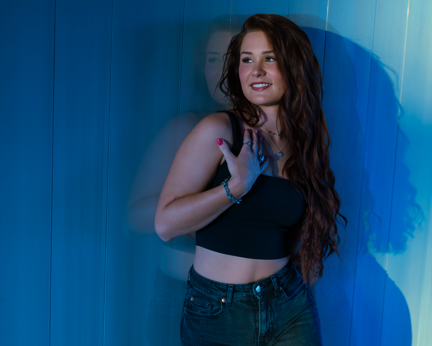 Young woman with long red hair posing against a blue wall in a black crop top and jeans.