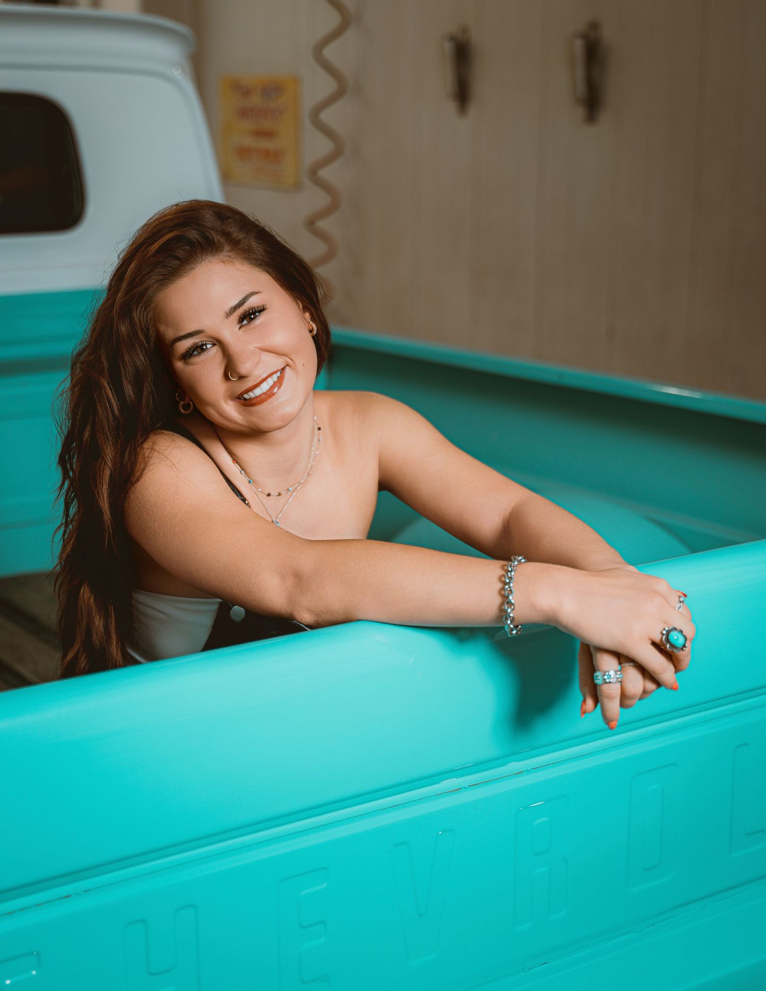 Smiling young woman with long brown hair posing in a turquoise truck bed, wearing silver jewelry.