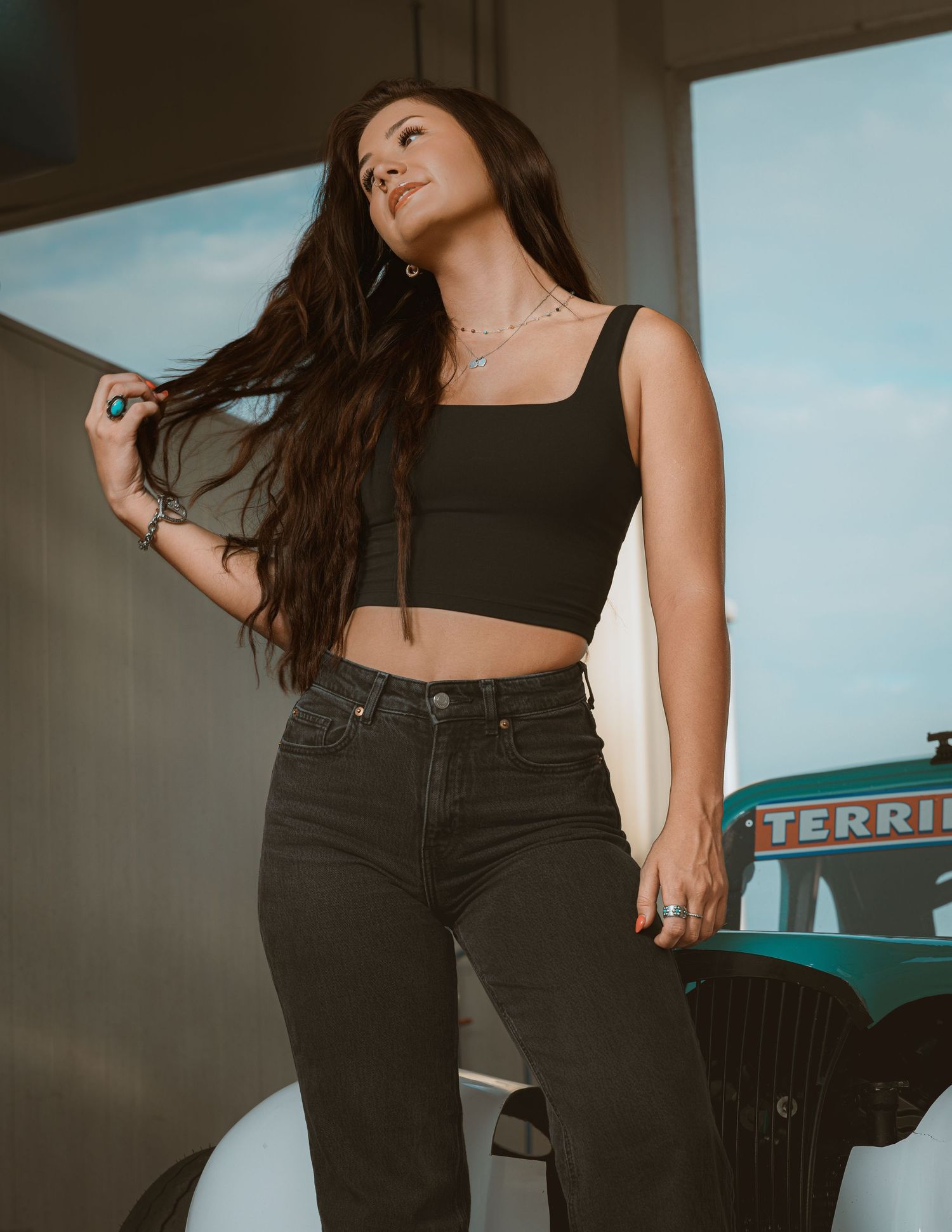 Stylish woman in black crop top and dark jeans holding her long hair, posing outdoors near a vintage teal vehicle.