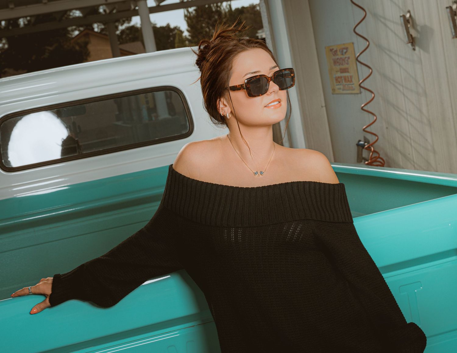 Woman in black off-shoulder sweater and sunglasses posing against a turquoise vintage truck outdoors.