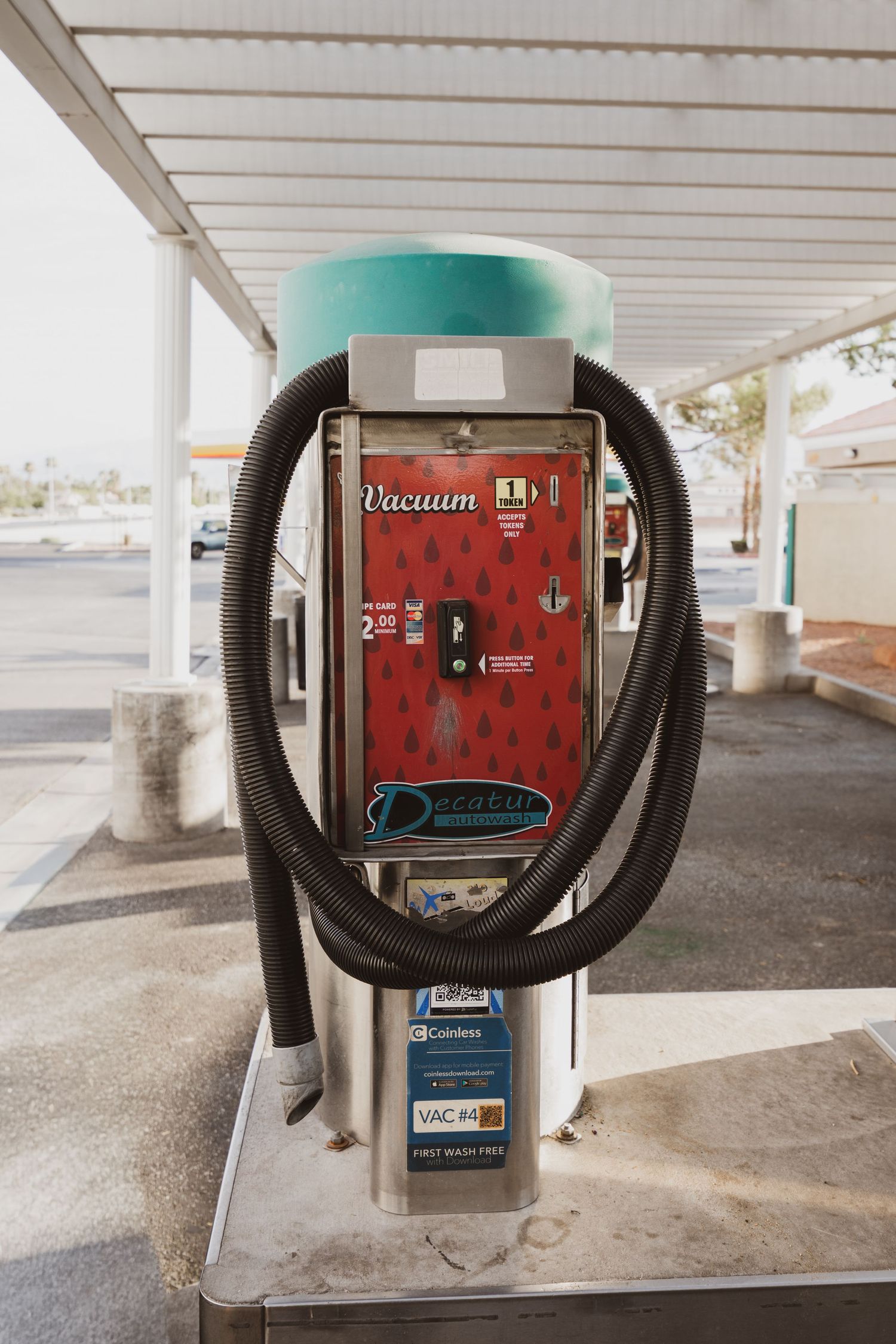 Red coin-operated vacuum station with hose at a self-service car wash facility under a covered canopy.