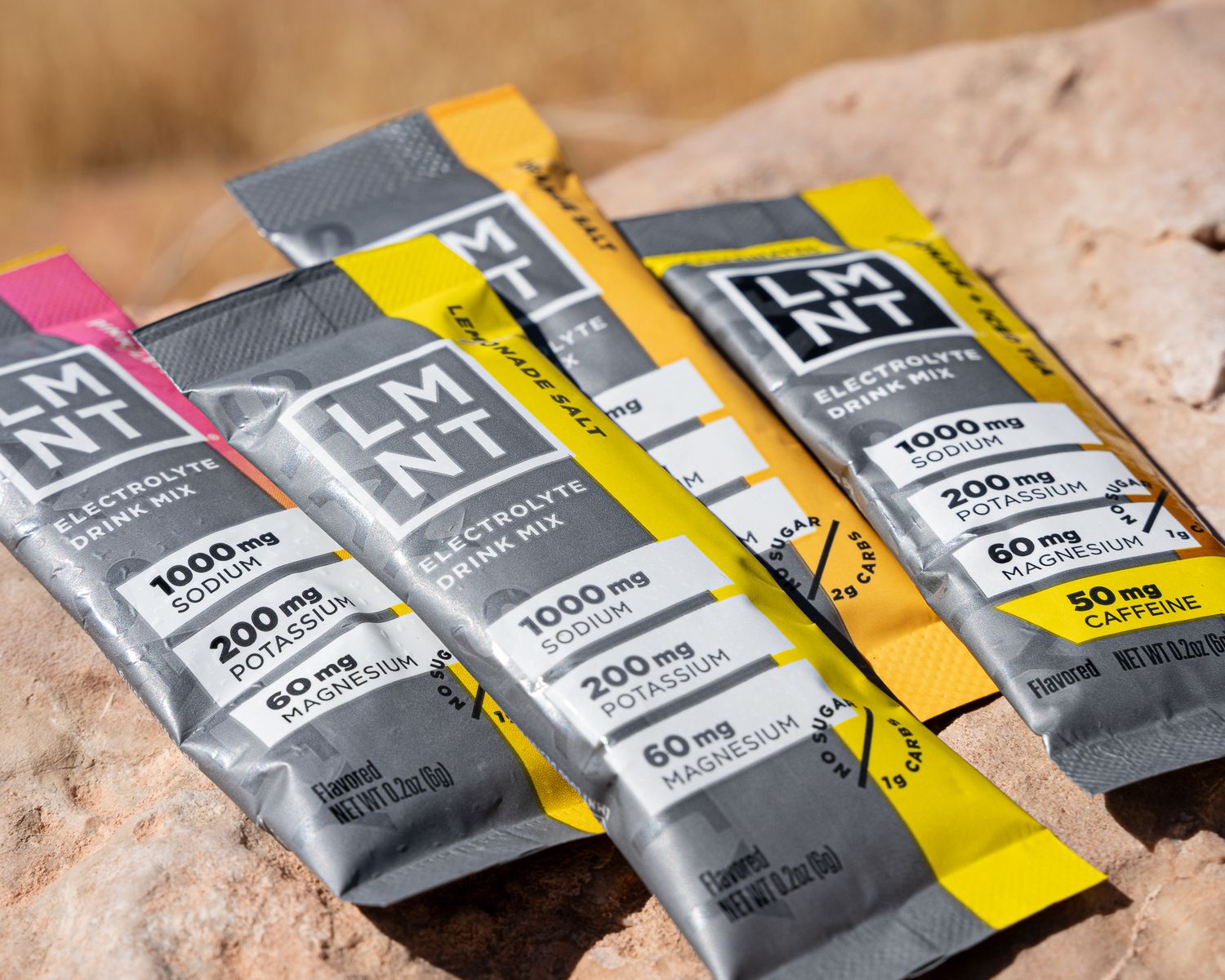 Assorted LMNT electrolyte drink mix packets in pink, yellow, and gray on rocky surface.
