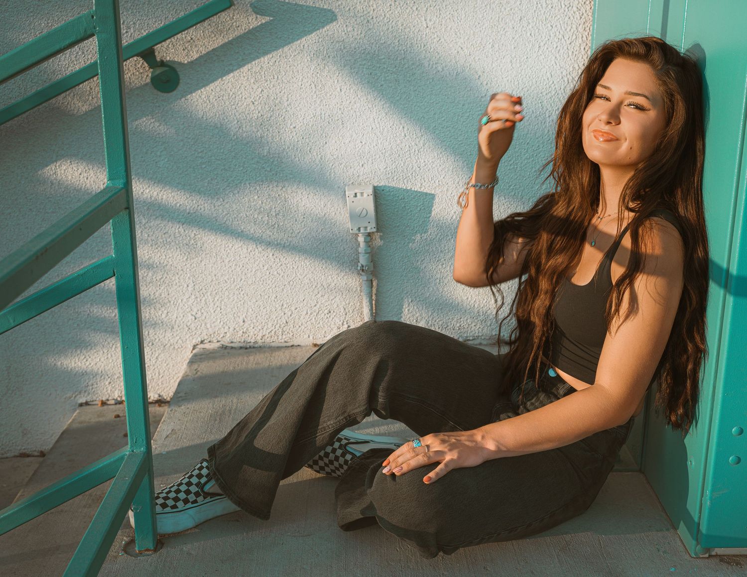 Young woman with long brown hair sitting on ground against white wall near teal staircase, holding a small object.